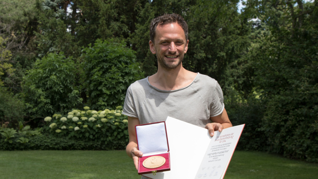 A person stands on a lawn, proudly holding a red case with a gold medal and an open certificate from the Adult Education Awards. They smile in a gray shirt, surrounded by lush green trees and bushes, embodying the spirit of learning celebrated by TM Wissen.