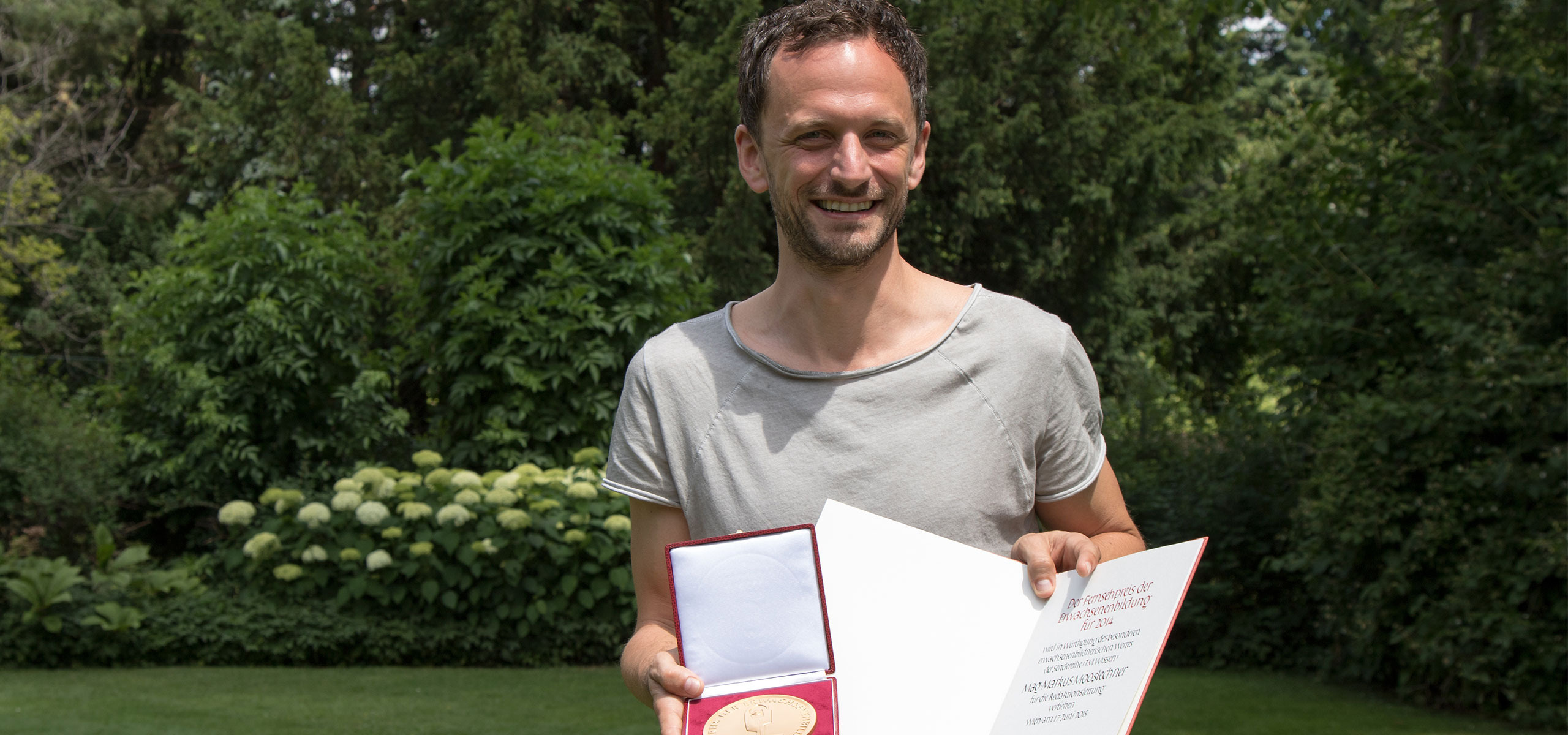 A person stands on a lawn, proudly holding a red case with a gold medal and an open certificate from the Adult Education Awards. They smile in a gray shirt, surrounded by lush green trees and bushes, embodying the spirit of learning celebrated by TM Wissen.