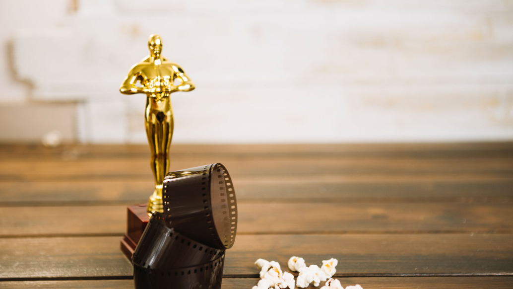 A golden trophy resembling an Oscar award statue stands on a wooden surface. Beside it, a coiled strip of film and some scattered popcorn are visible. The background is softly blurred.