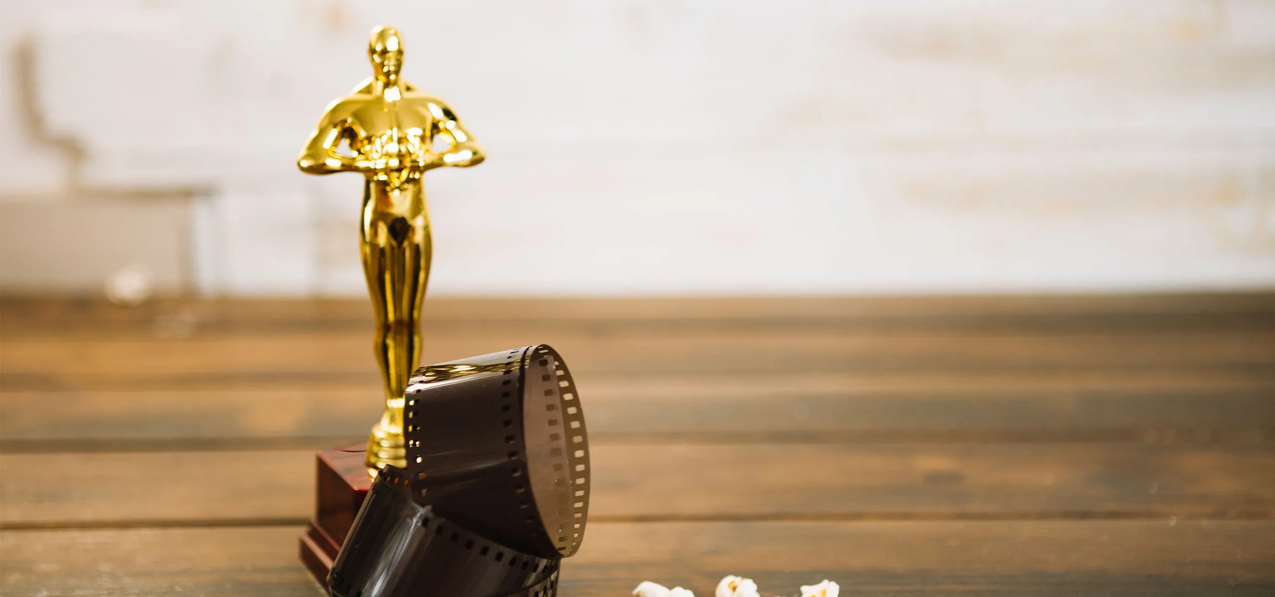 A golden trophy resembling an Oscar award statue stands on a wooden surface. Beside it, a coiled strip of film and some scattered popcorn are visible. The background is softly blurred.