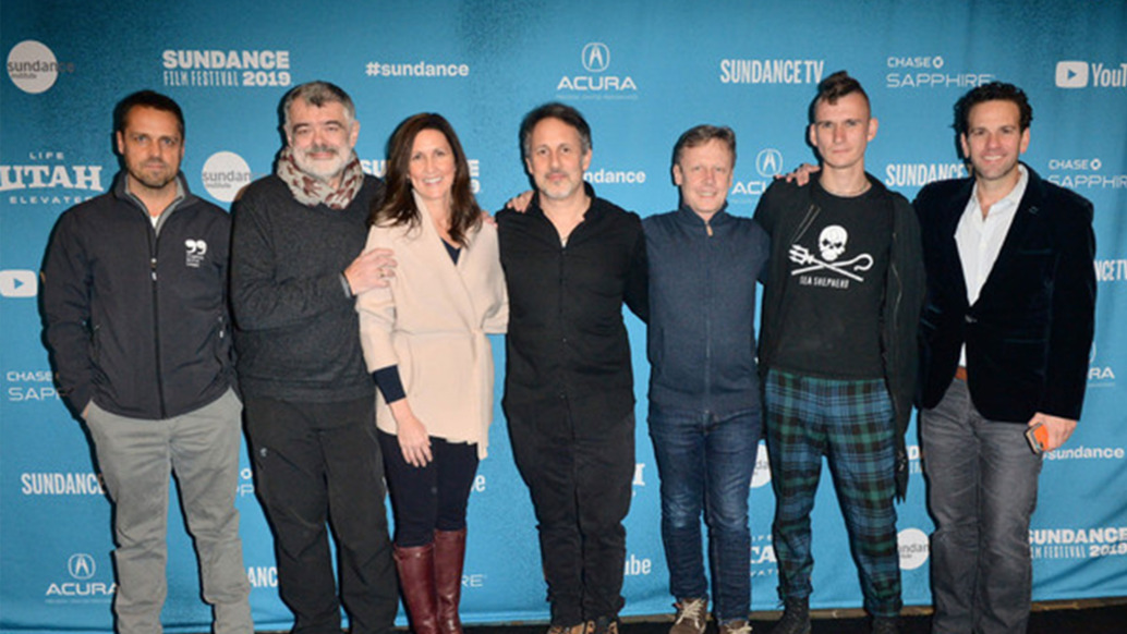 A group of seven people stands in front of a blue backdrop adorned with Sundance Film Festival 2019 and sponsor logos, celebrating the Sundance Premiere of "Sea of Shadows," as they pose and smile for the camera.