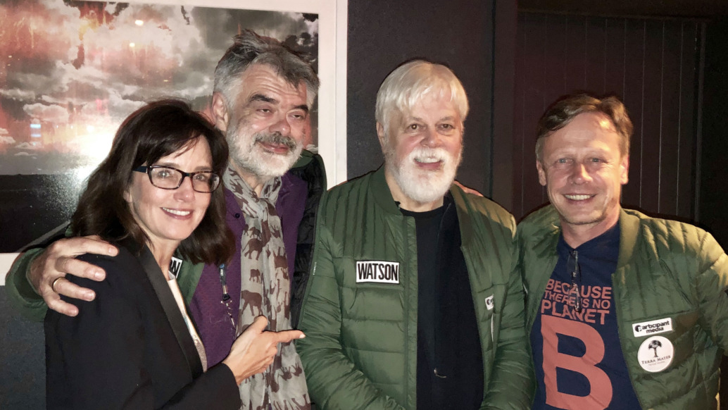 Four people are standing together indoors, smiling at the camera. Three are wearing green jackets, one with a name tag reading Watson. The setting exudes an artistic vibe reminiscent of Tribeca Film Festival, with a dark-colored wall and a framed picture in the background.