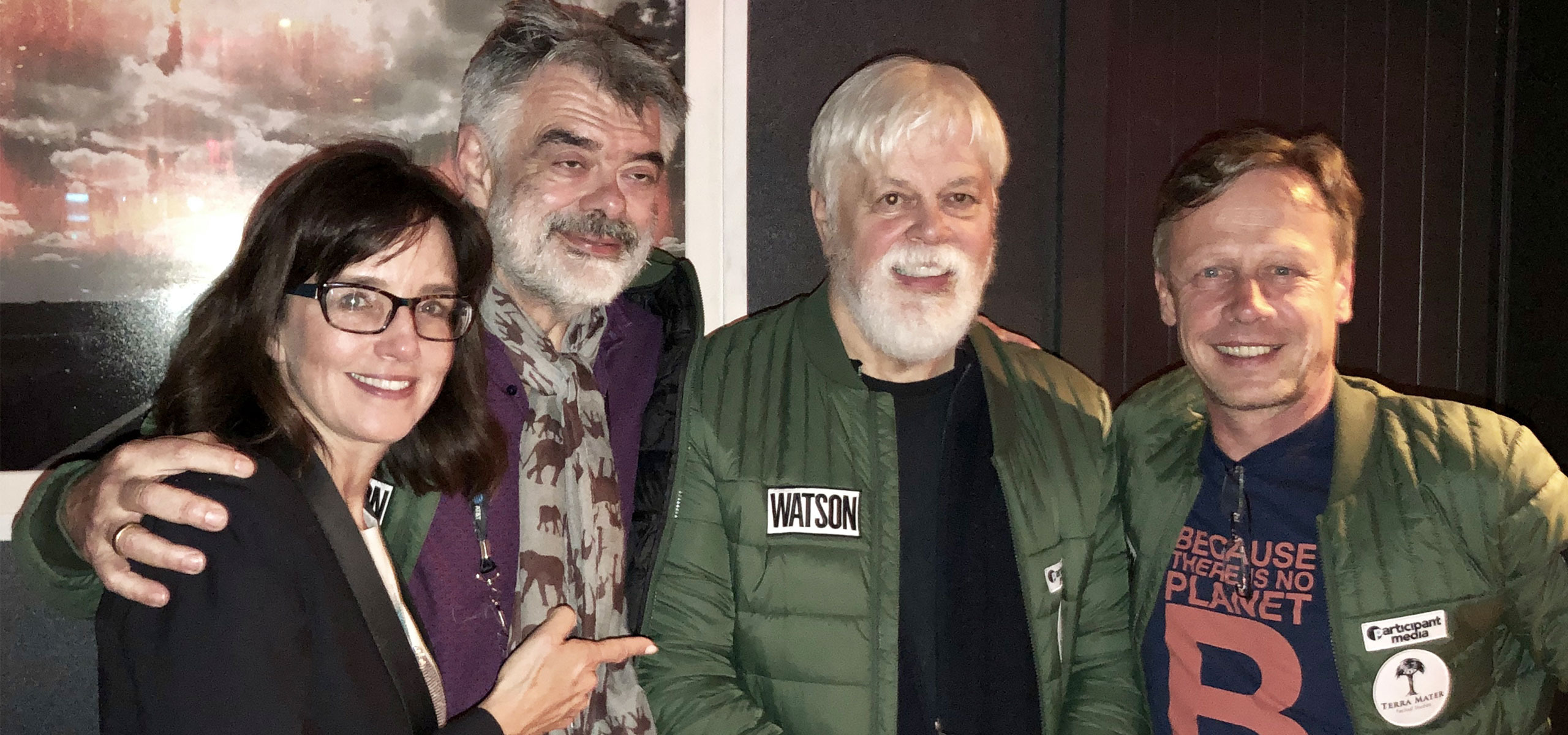 Four people are standing together indoors, smiling at the camera. Three are wearing green jackets, one with a name tag reading Watson. The setting exudes an artistic vibe reminiscent of Tribeca Film Festival, with a dark-colored wall and a framed picture in the background.