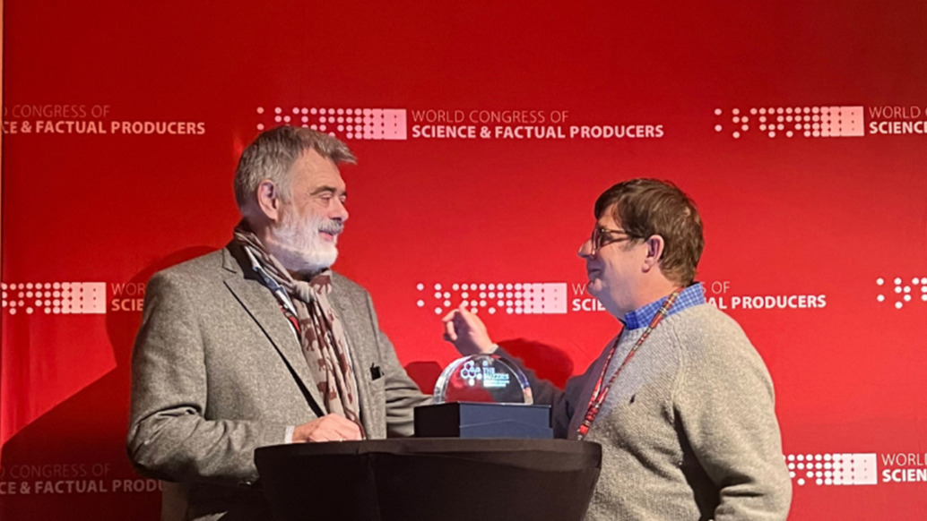 Two men stand on a stage in front of a red backdrop that reads World Congress of Science & Factual Producers. One man gestures toward the other, who is about to receive the Lifetime Achievement Award. A small trophy sits on a table between them, marking this WCSFP 2022 moment.