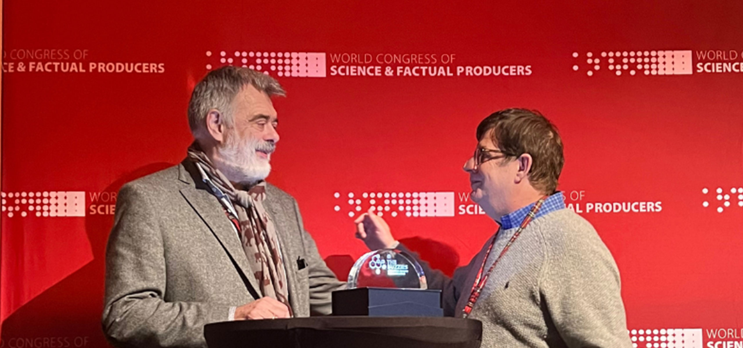 Two men stand on a stage in front of a red backdrop that reads World Congress of Science & Factual Producers. One man gestures toward the other, who is about to receive the Lifetime Achievement Award. A small trophy sits on a table between them, marking this WCSFP 2022 moment.