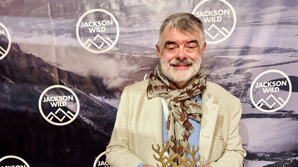 An older man with a gray beard, wearing a cream jacket and patterned scarf, proudly holds an award at the Jackson Wild Media Awards backdrop, adorned with logos and a mountain scene.