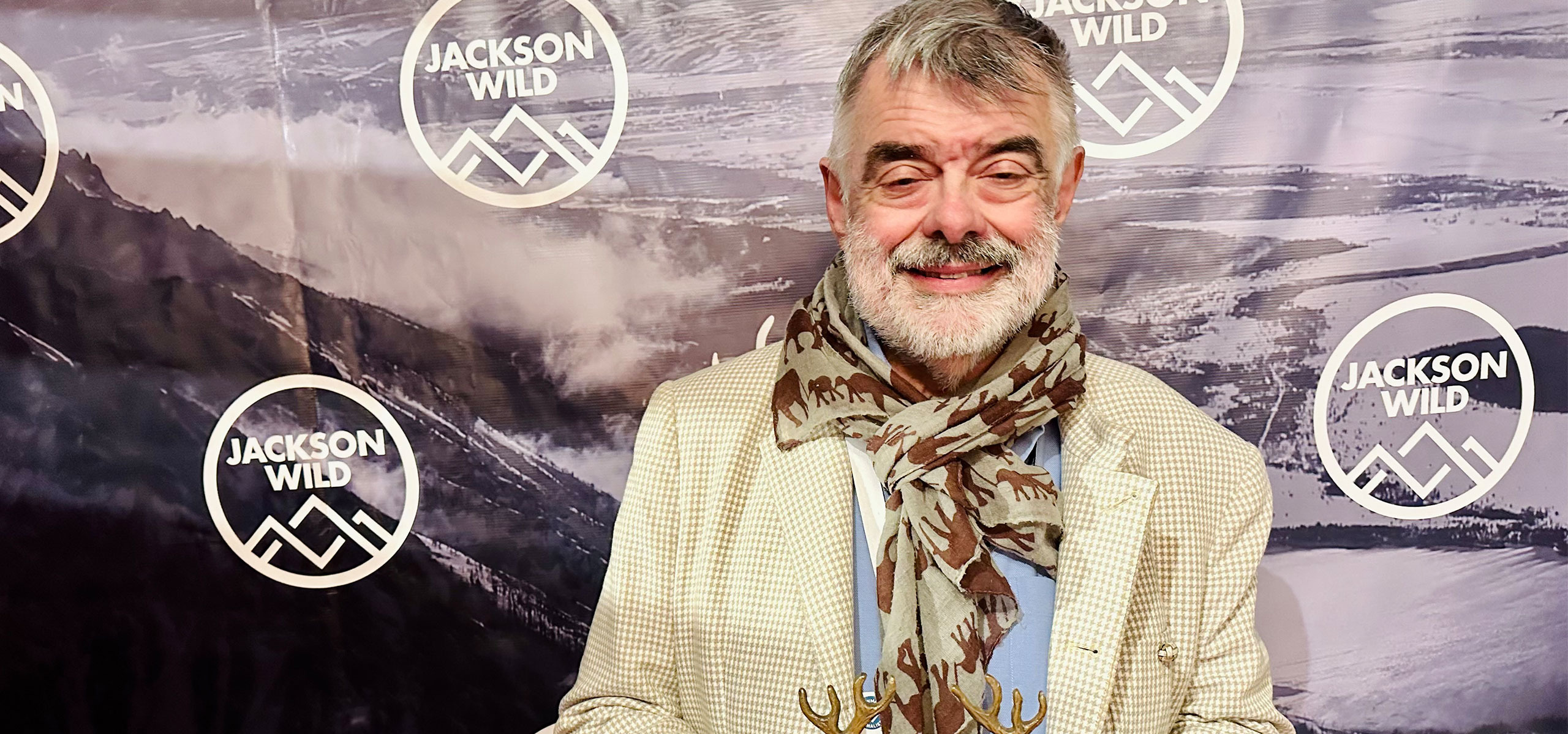 An older man with a gray beard, wearing a cream jacket and patterned scarf, proudly holds an award at the Jackson Wild Media Awards backdrop, adorned with logos and a mountain scene.