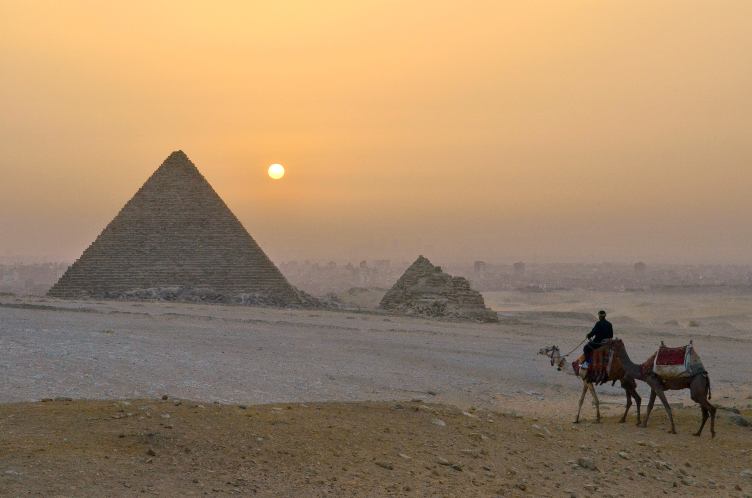 As the sun dips below the horizon, a person rides a camel through the sandy landscape, with two pyramids towering in the distance under a hazy sky, reminiscent of tales along the banks of the Nile.