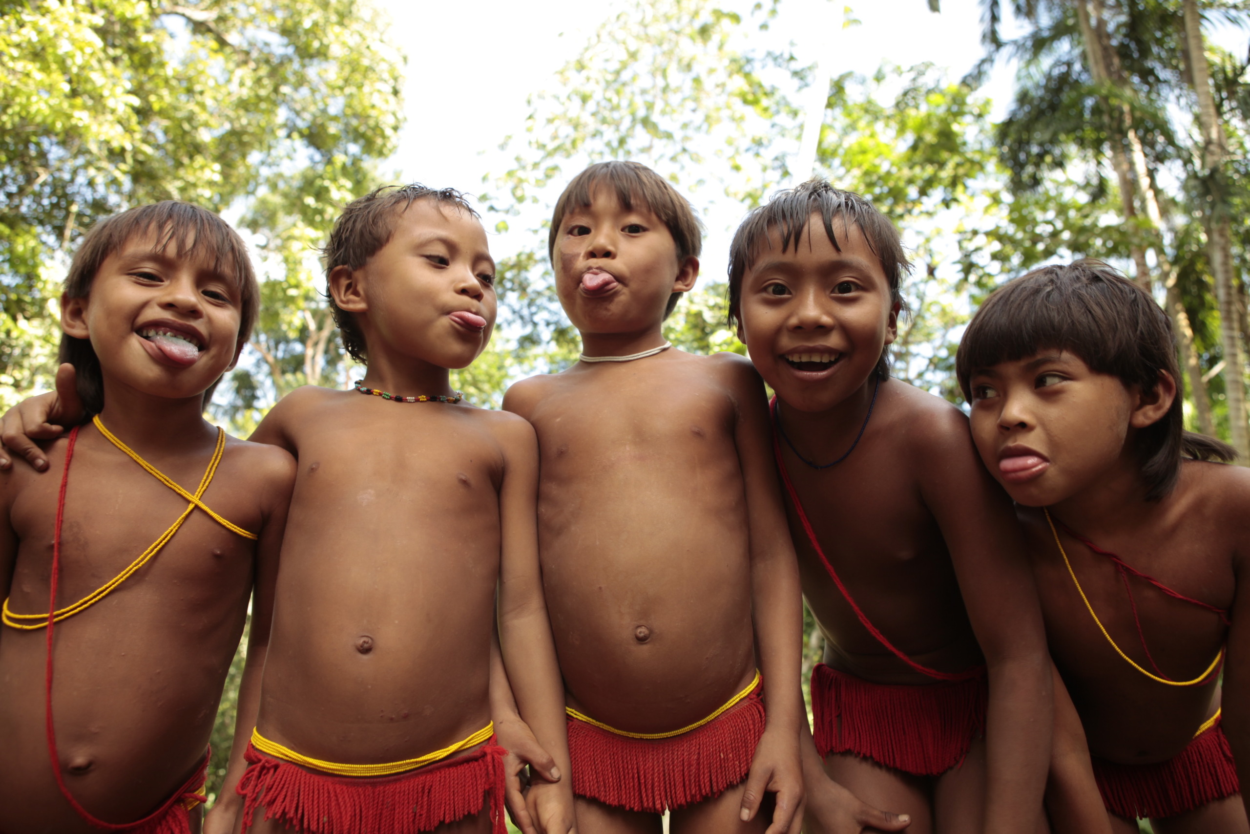 Five children wearing red traditional attire stand in a forest setting, smiling and playfully sticking out their tongues. They seem between worlds, surrounded by lush green trees that suggest a vibrant, natural environment.