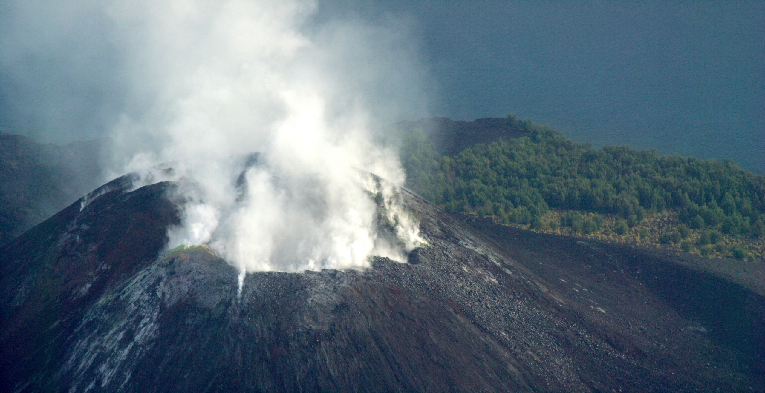 Aerial view of a volcanic crater emitting white smoke on a remote island, surrounded by dark rocky terrain. In the background, a body of water and a patch of green vegetation are visible under a cloudy sky, as if time stands still in this rugged landscape.