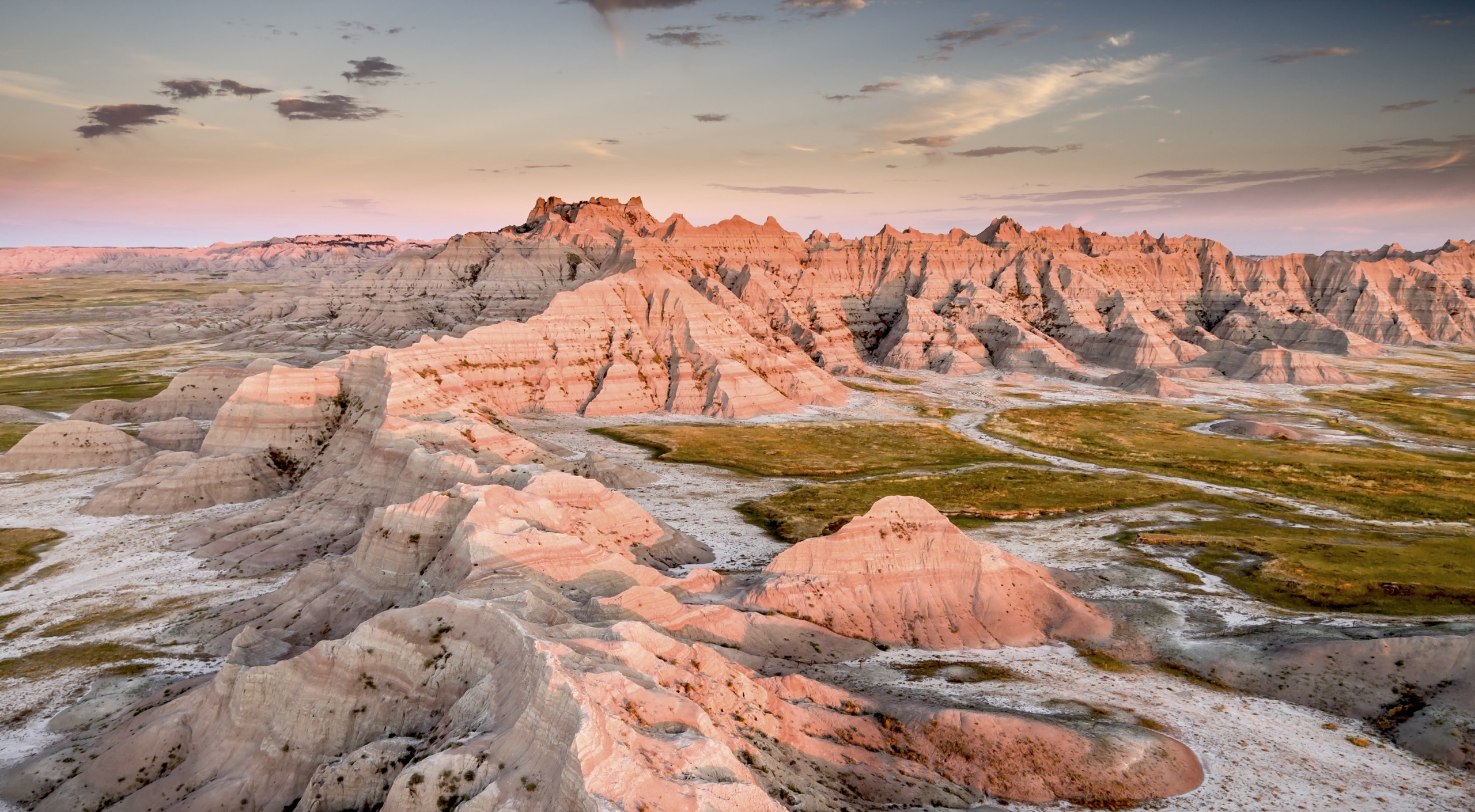 A stunning landscape of the Badlands at sunrise reveals the jagged rock formations and deep ravines illuminated in shades of pink and orange, contrasting with the lush green grasslands. A partly cloudy sky adds depth to this breathtaking scene in the Badlands.