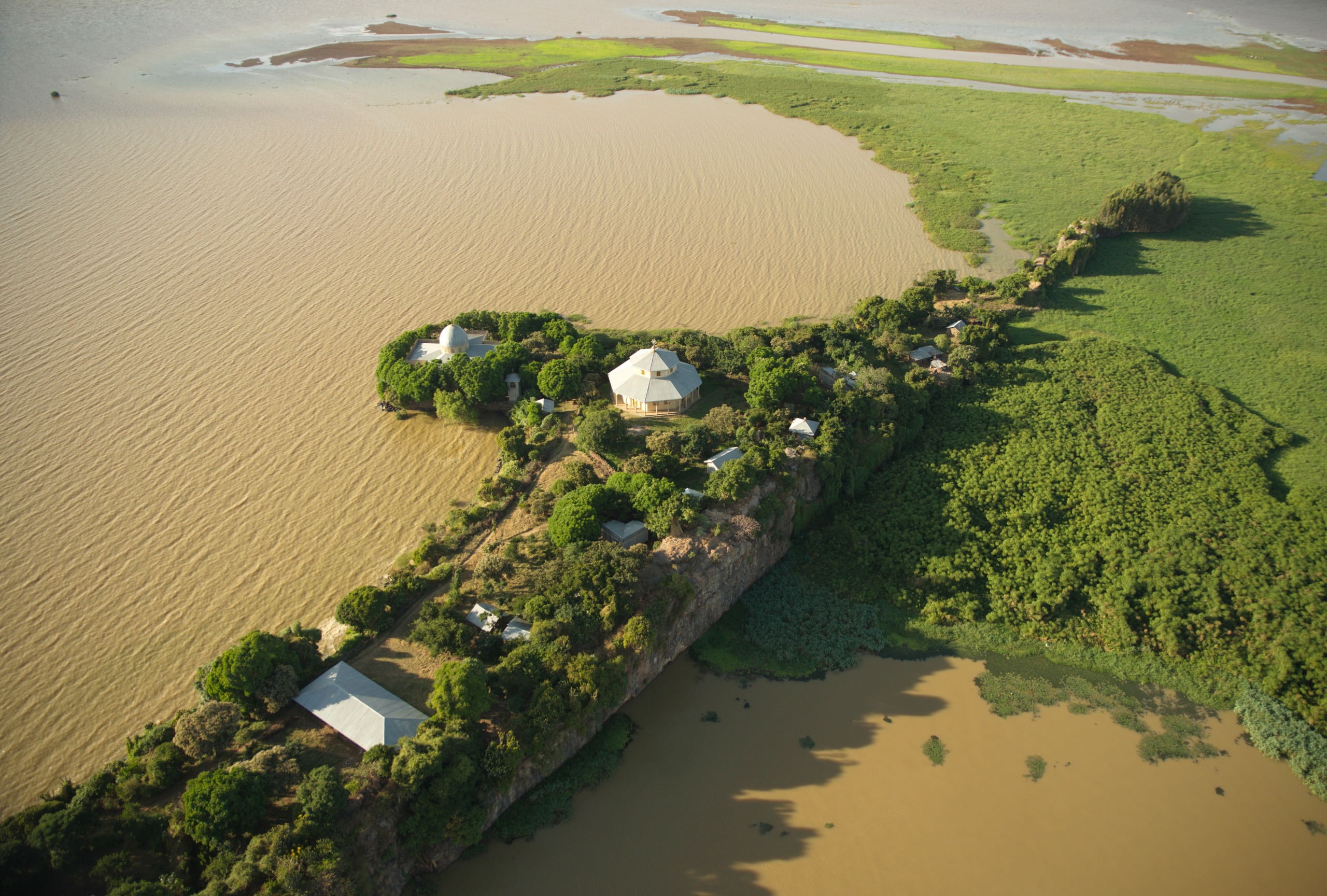 An aerial view reveals a lush, green island in the Nile, surrounded by muddy brown waters. Small buildings peek from among the trees, and a strip of greenery connects the island to the larger landmass, offering a serene yet vibrant landscape.