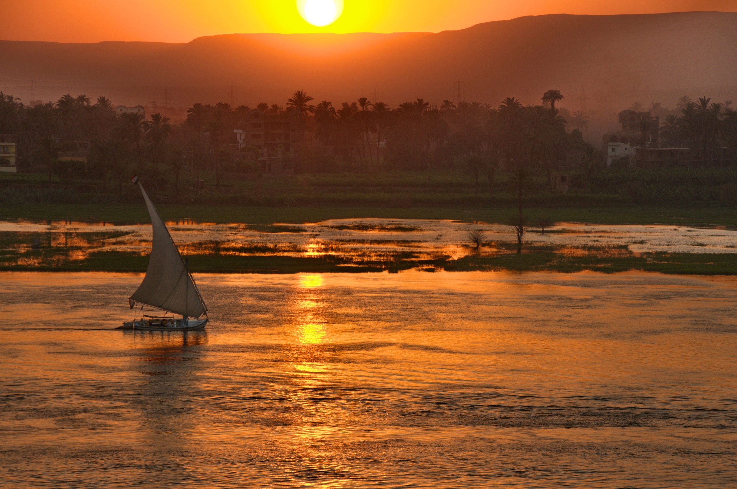 A sailboat glides across a tranquil river at sunset, reminiscent of the Nile, surrounded by lush greenery and silhouetted trees. The vibrant orange and yellow sky reflects on the water's surface, creating a warm, serene atmosphere.