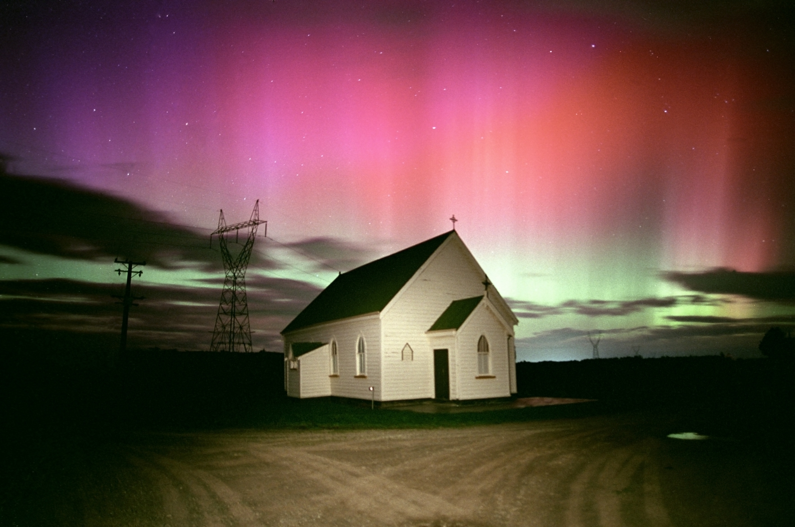 A small, white church at night rests under a sky illuminated with an aurora of vibrant pink and green. The silhouette of a power line intersects the scene, enhancing the serene yet dramatic atmosphere.