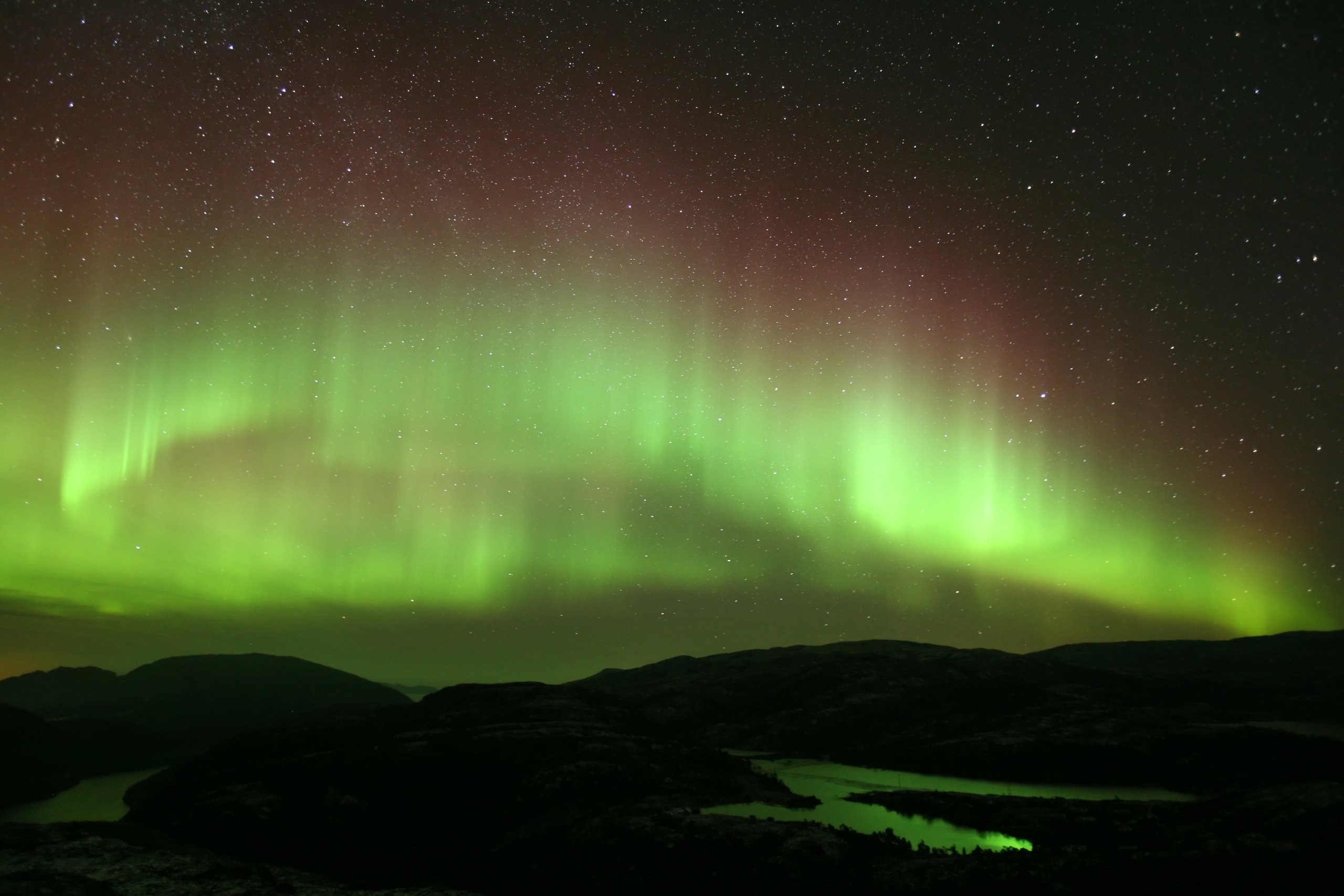 A night sky illuminated by vibrant auroras in green and yellow dances over a dark, silhouetted landscape with hills and reflective water bodies, sparkling under a starry sky.