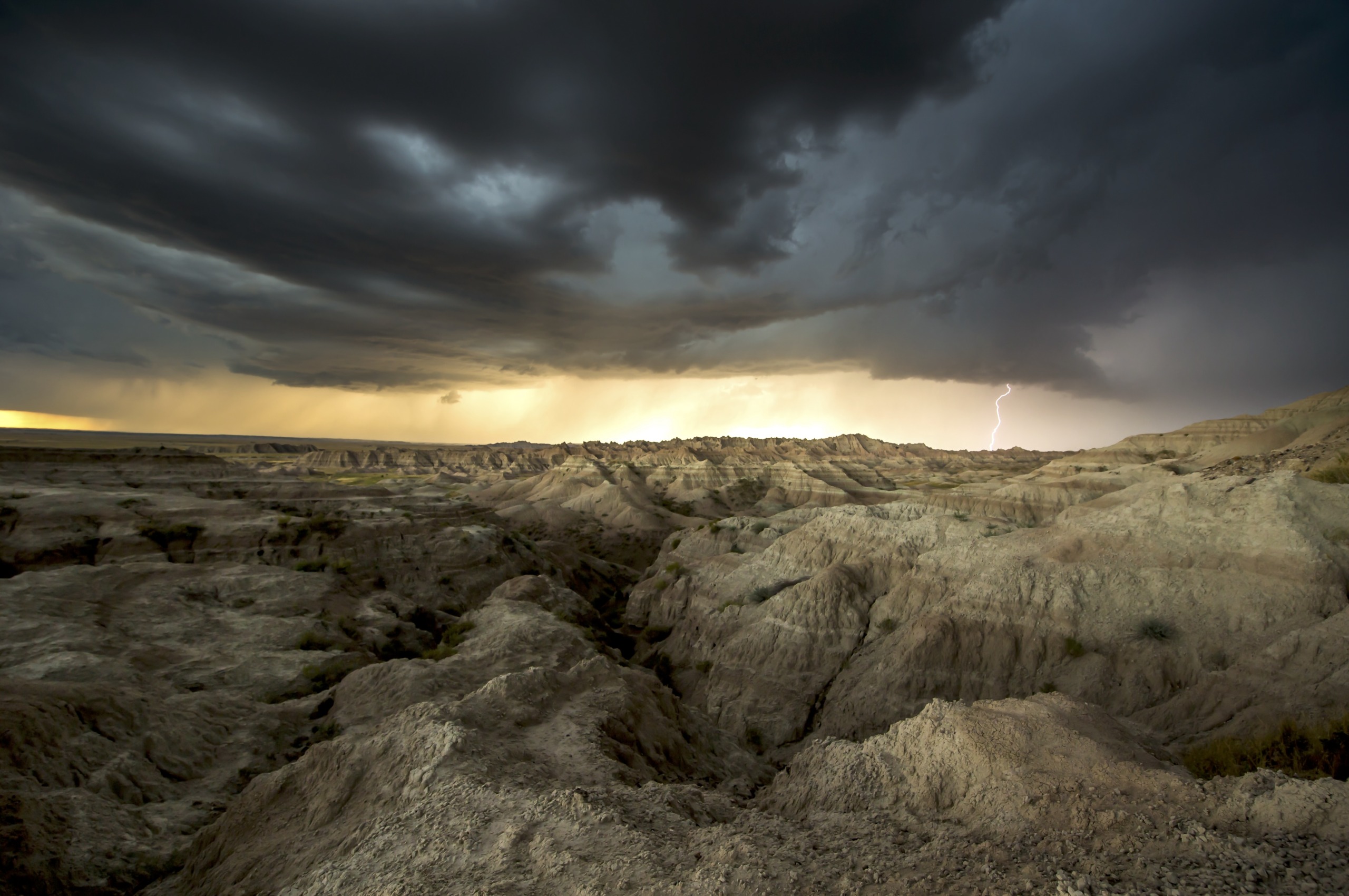 A dramatic landscape of rugged, rocky terrain resembles the badlands under a dark, stormy sky. Lightning is visible in the distance, illuminating part of the horizon. The scene conveys a sense of natural power and vastness.