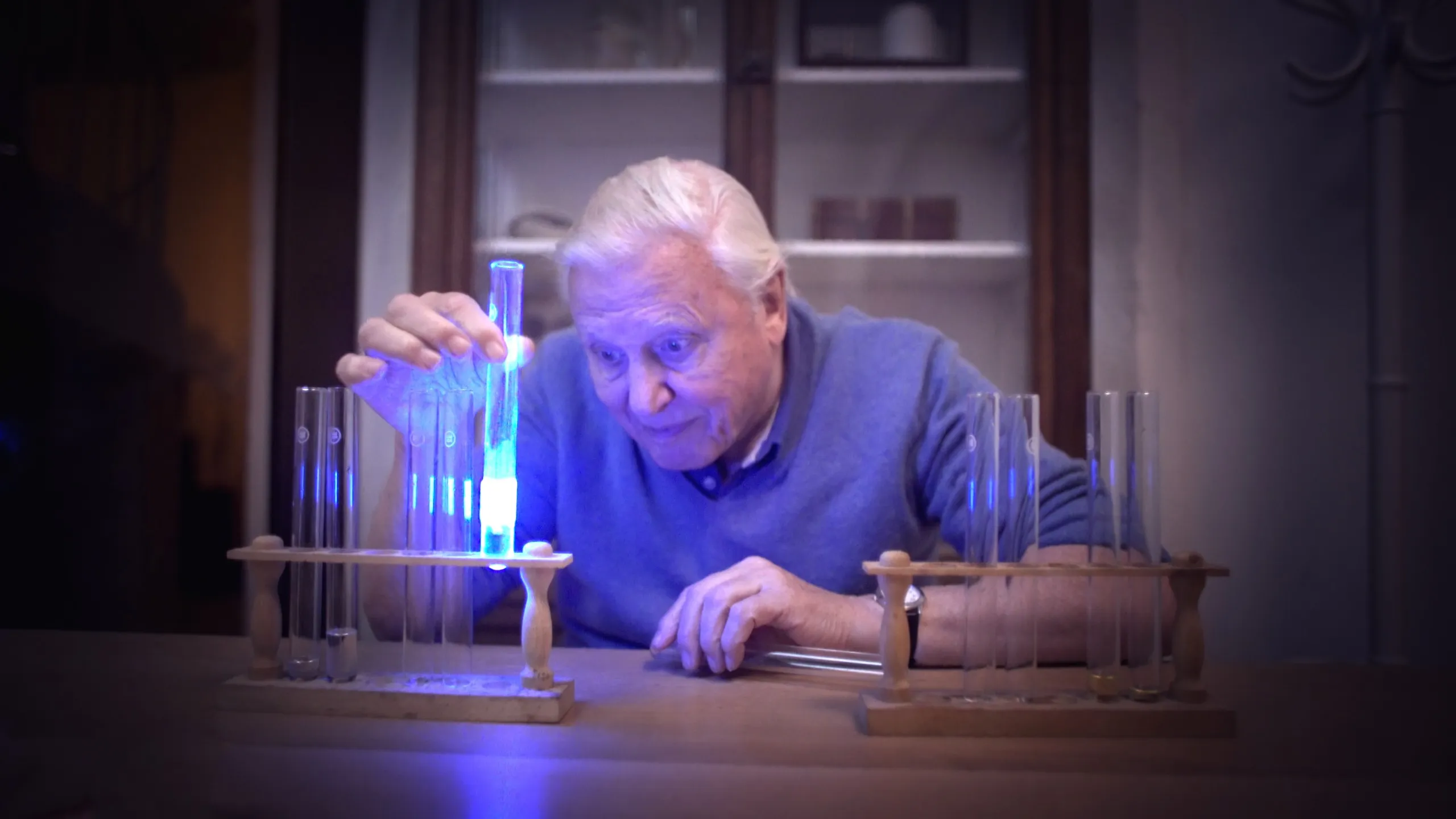 An older man with white hair inspects a glowing blue liquid in a test tube, reminiscent of scenes from a David Attenborough documentary. Sitting at a table with several other test tubes in wooden stands, the dimly lit room emphasizes the ethereal blue glow of this light on Earth.