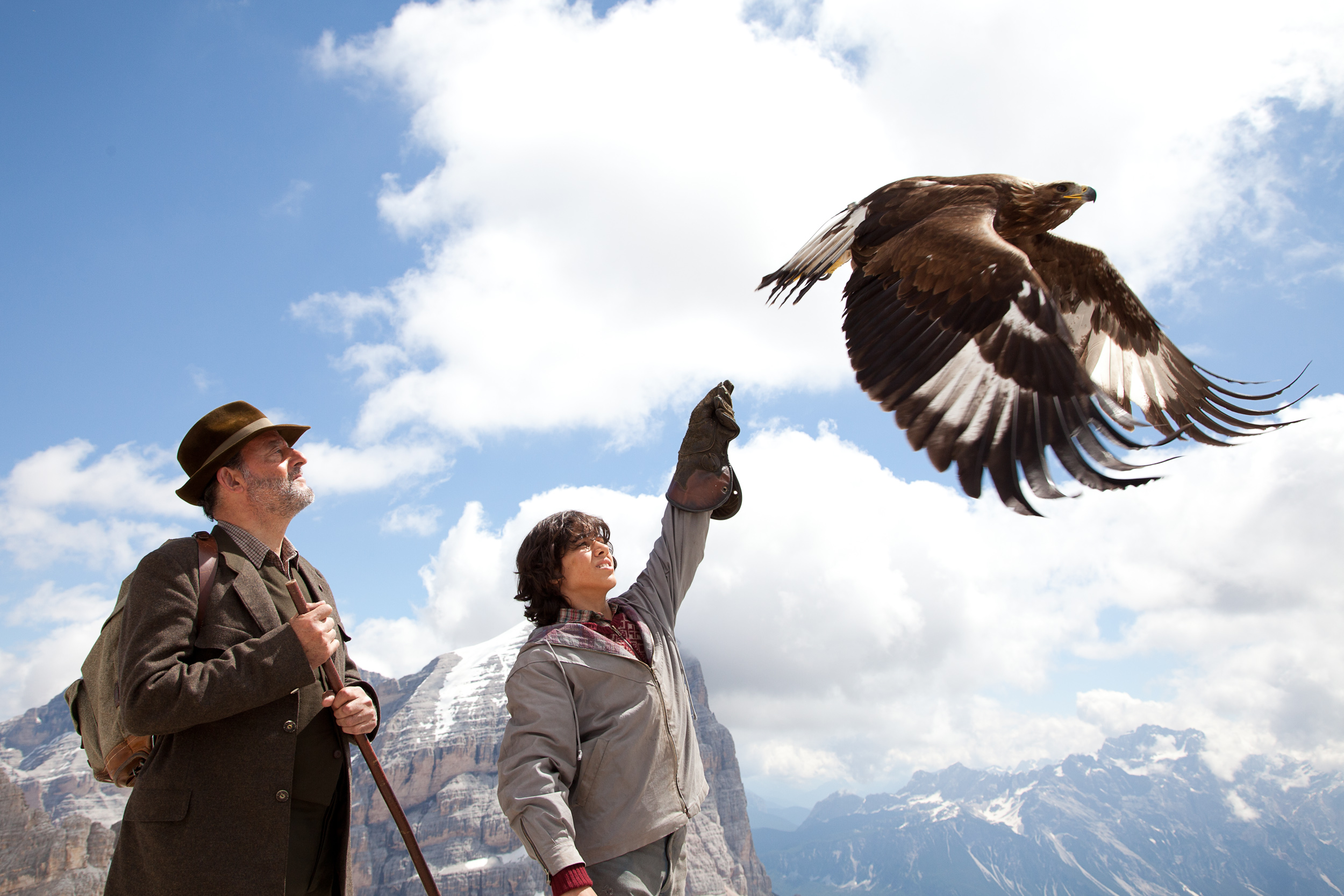 In a mountainous landscape under partly cloudy skies, a person releases a large eagle into the sky as another looks on. Both dressed in outdoor clothing, they resemble Brothers of the Wind, united with nature's majesty.