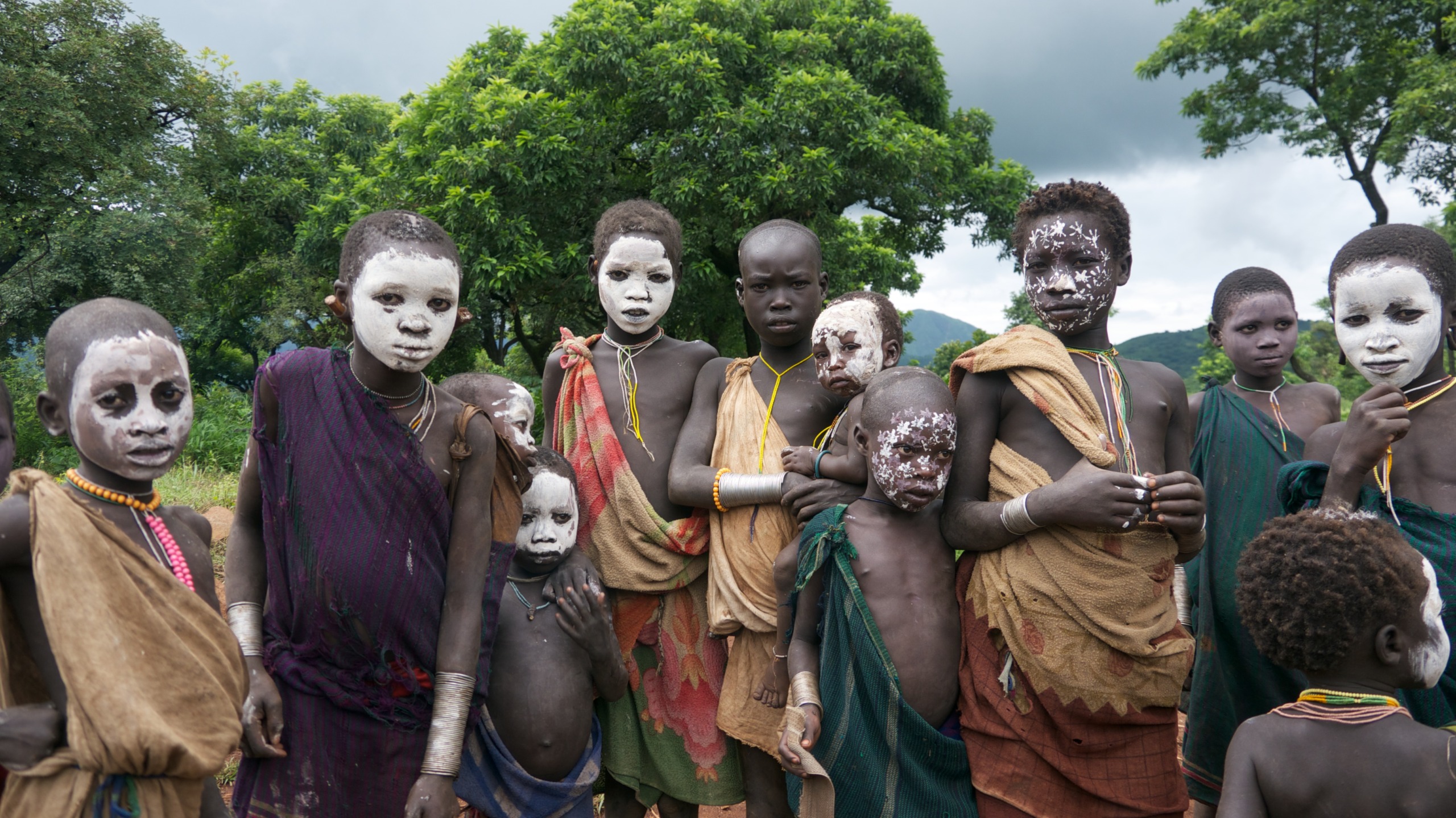 A group of people, including children, stand outdoors in traditional attire, their faces painted with white patterns. Lush green trees and an overcast sky create a backdrop that suggests a realm between worlds, merging the natural setting with an ethereal atmosphere.