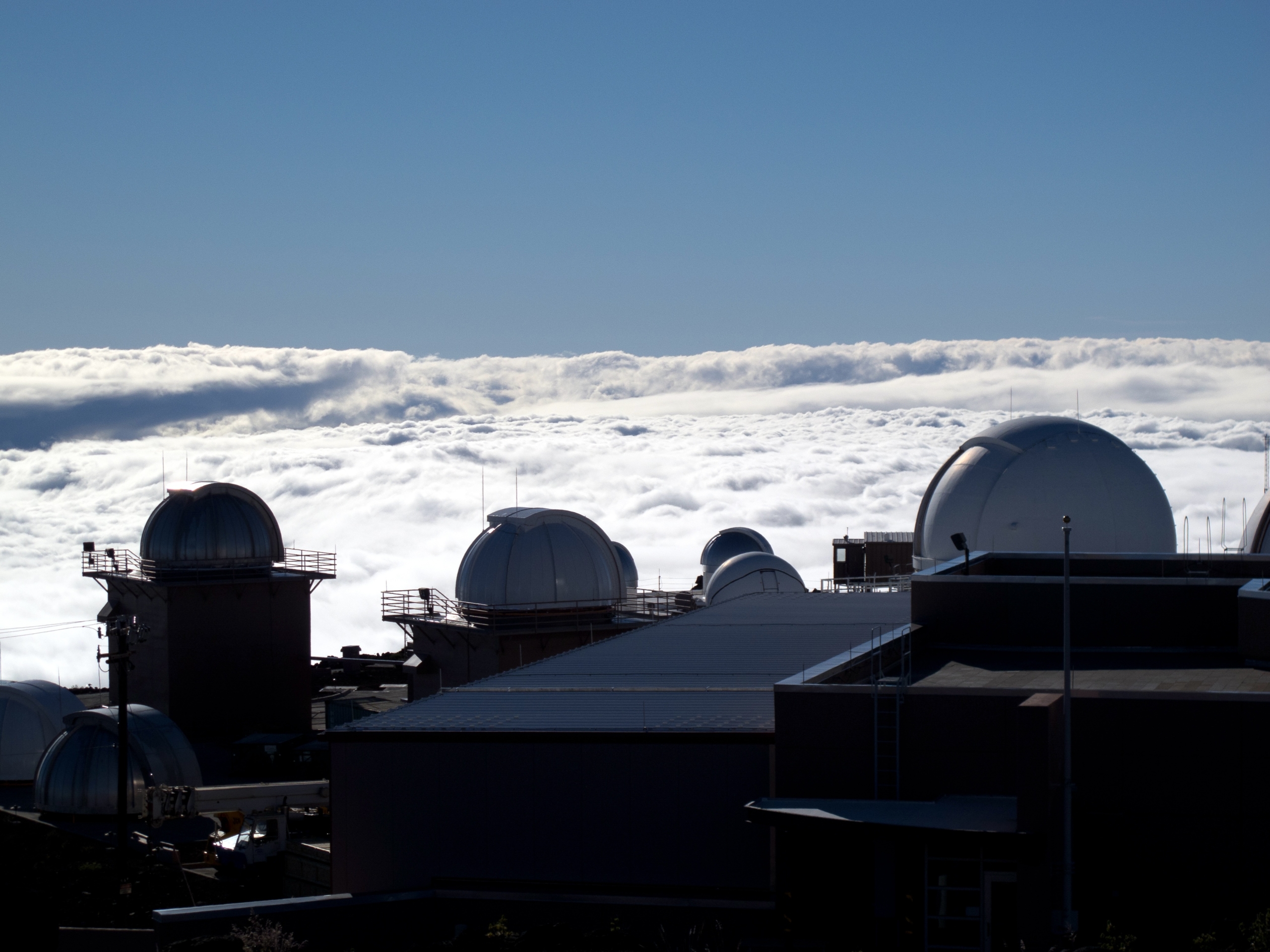 A cluster of observatory domes sits against a backdrop of a bright blue sky and thick white clouds. Some domes are open, revealing telescopes used for meteor and astronomical observations. The scene exudes a peaceful, scientific atmosphere.