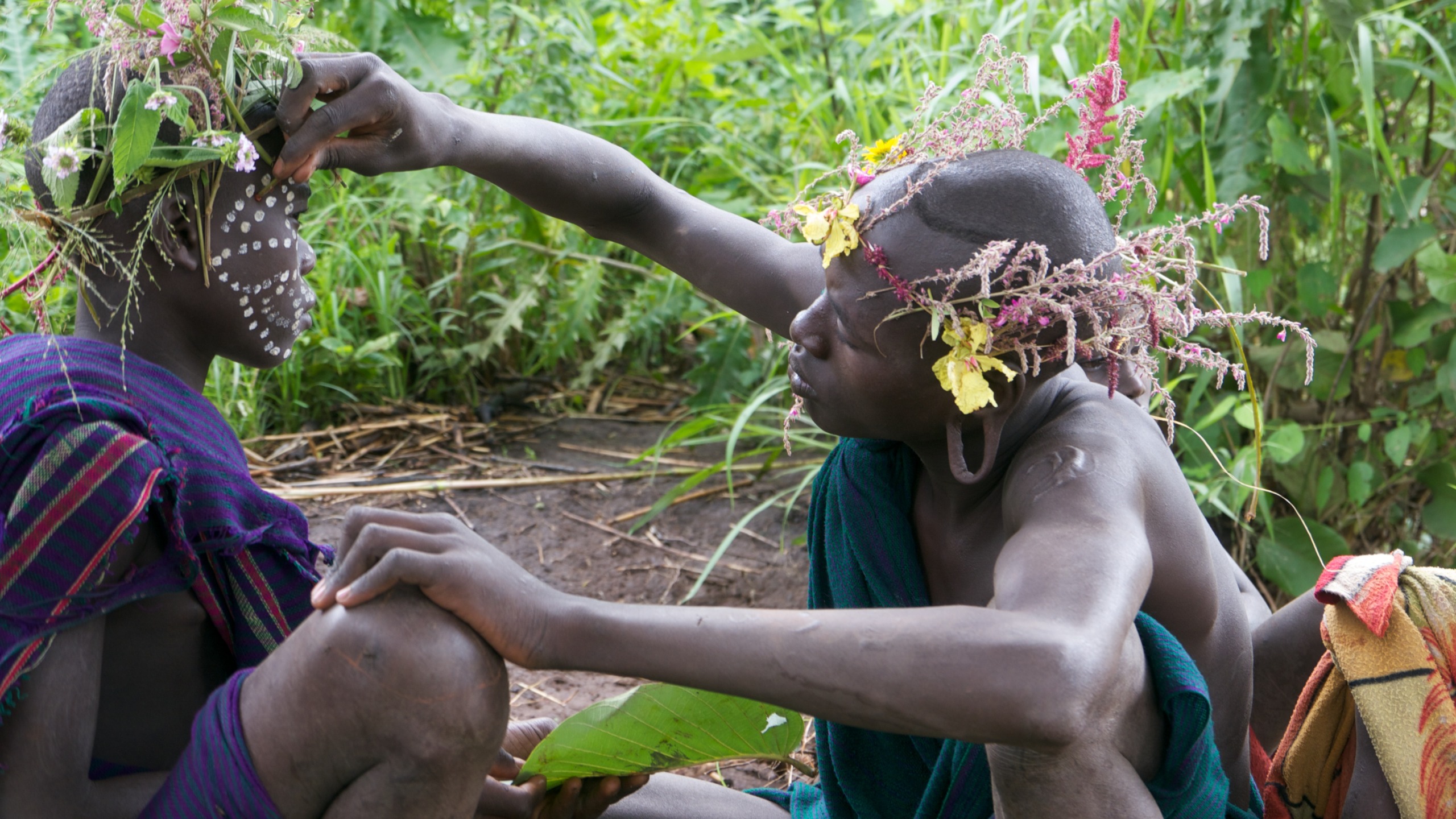 Two people with floral headdresses sit outdoors, surrounded by lush greenery—between worlds of tradition and nature. One person applies white face paint with intricate dots and lines to the other's face. Both wear traditional garments, creating a vibrant, natural scene seamlessly blending cultures.