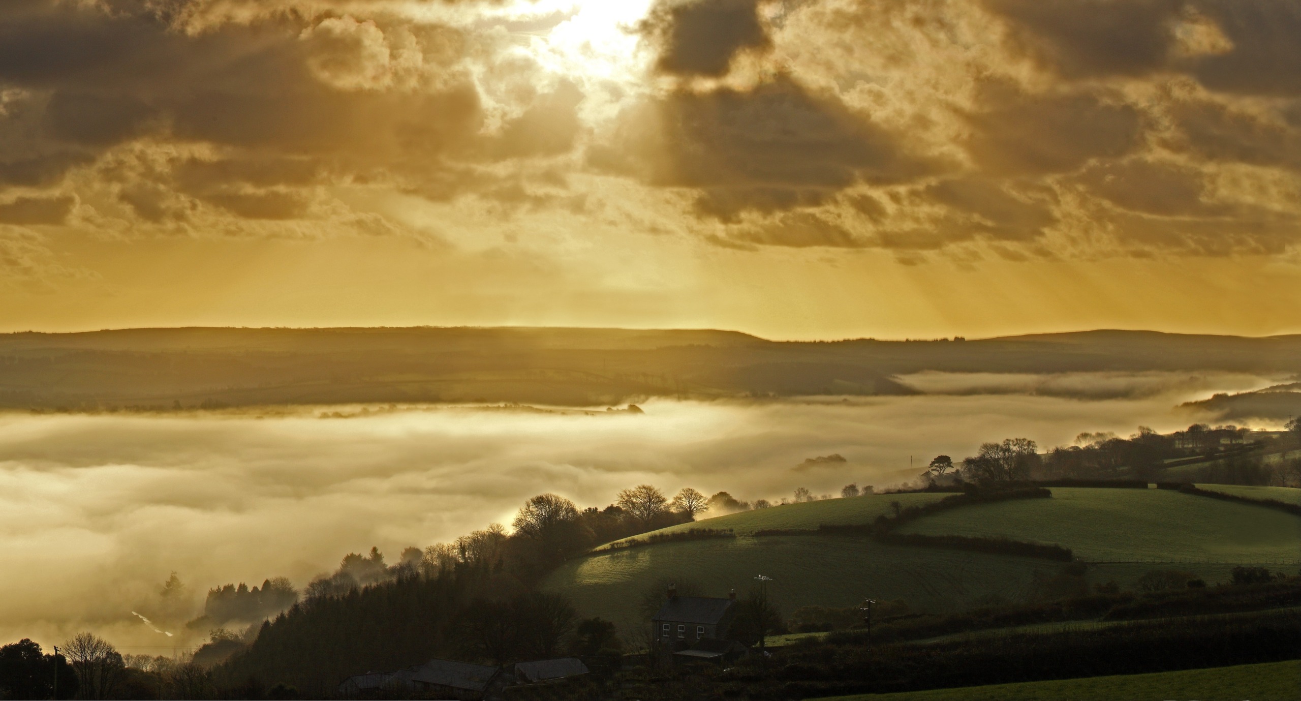 Sunlight breaks through dramatic clouds, casting a golden glow over Cornwall's lush green landscape. Fog rolls over the hills, partially obscuring the valleys, while trees and a lone building are visible in the foreground, enhancing the region's ethereal beauty.