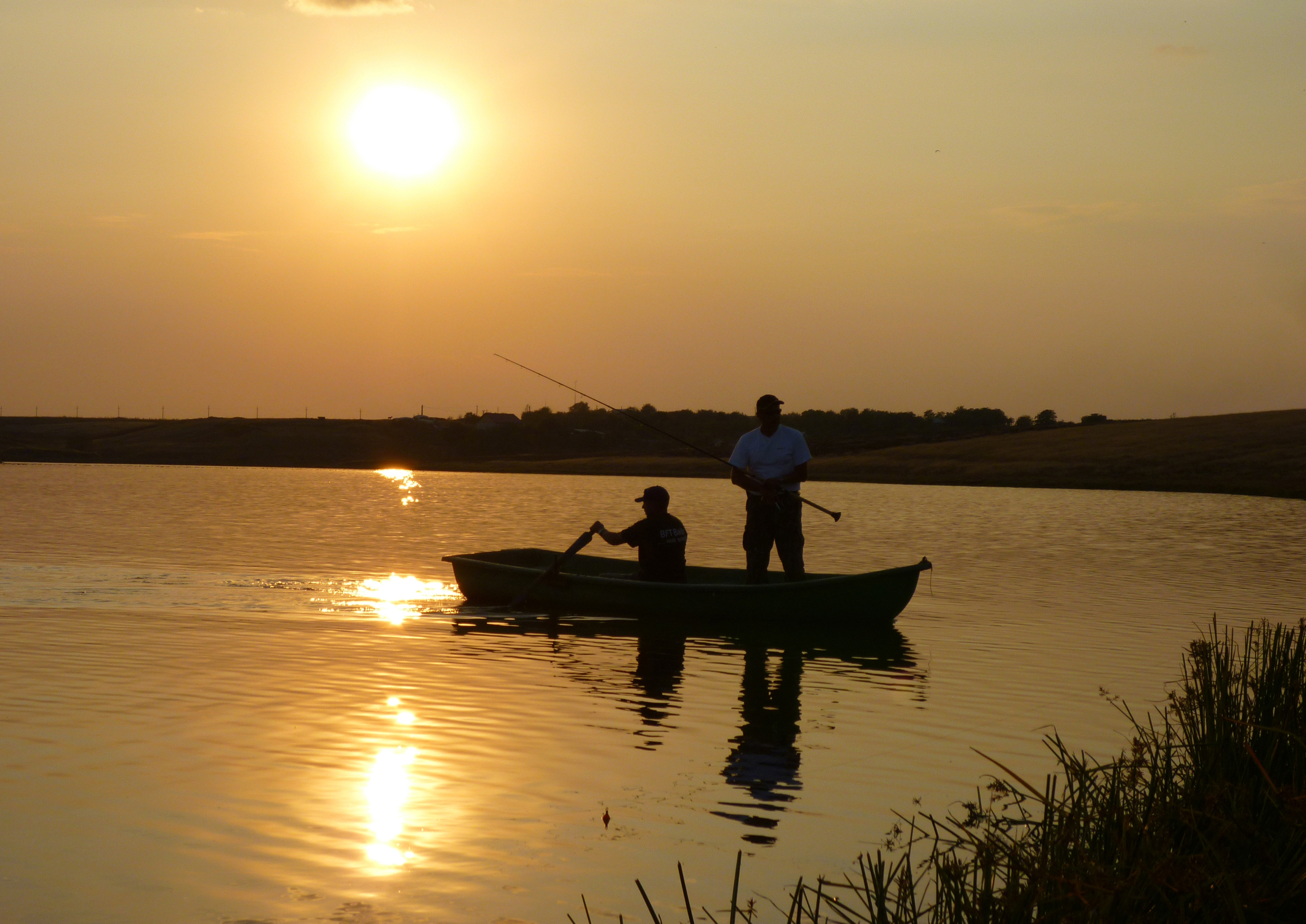 Two people in a rowboat fish on a calm lake at sunset, where the sun casts a golden reflection on the water, creating a serene and peaceful atmosphere. Nearby, beluga-shaped clouds drift lazily across the sky, while tall grasses sway gently in the foreground.