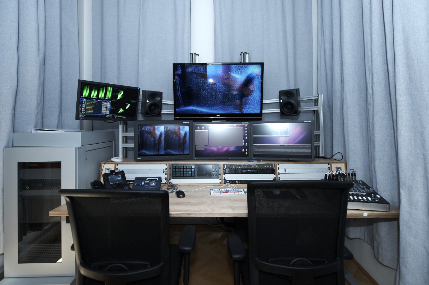 A modern audio-visual production studio with multiple screens displaying video content. Two black chairs face the workstation, which includes mixing equipment, speakers, and a control panel. Grey curtains hang in the background.