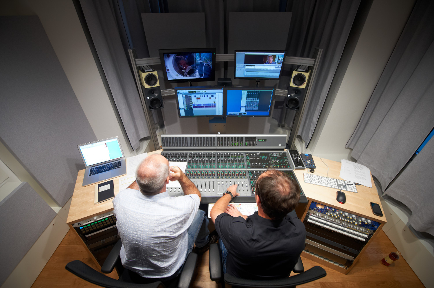 Two men sit in a recording studio, working at a large audio mixing console. Multiple screens display sound and video editing software. Papers, laptops, and speakers are arranged around the desk, creating a busy, focused atmosphere.