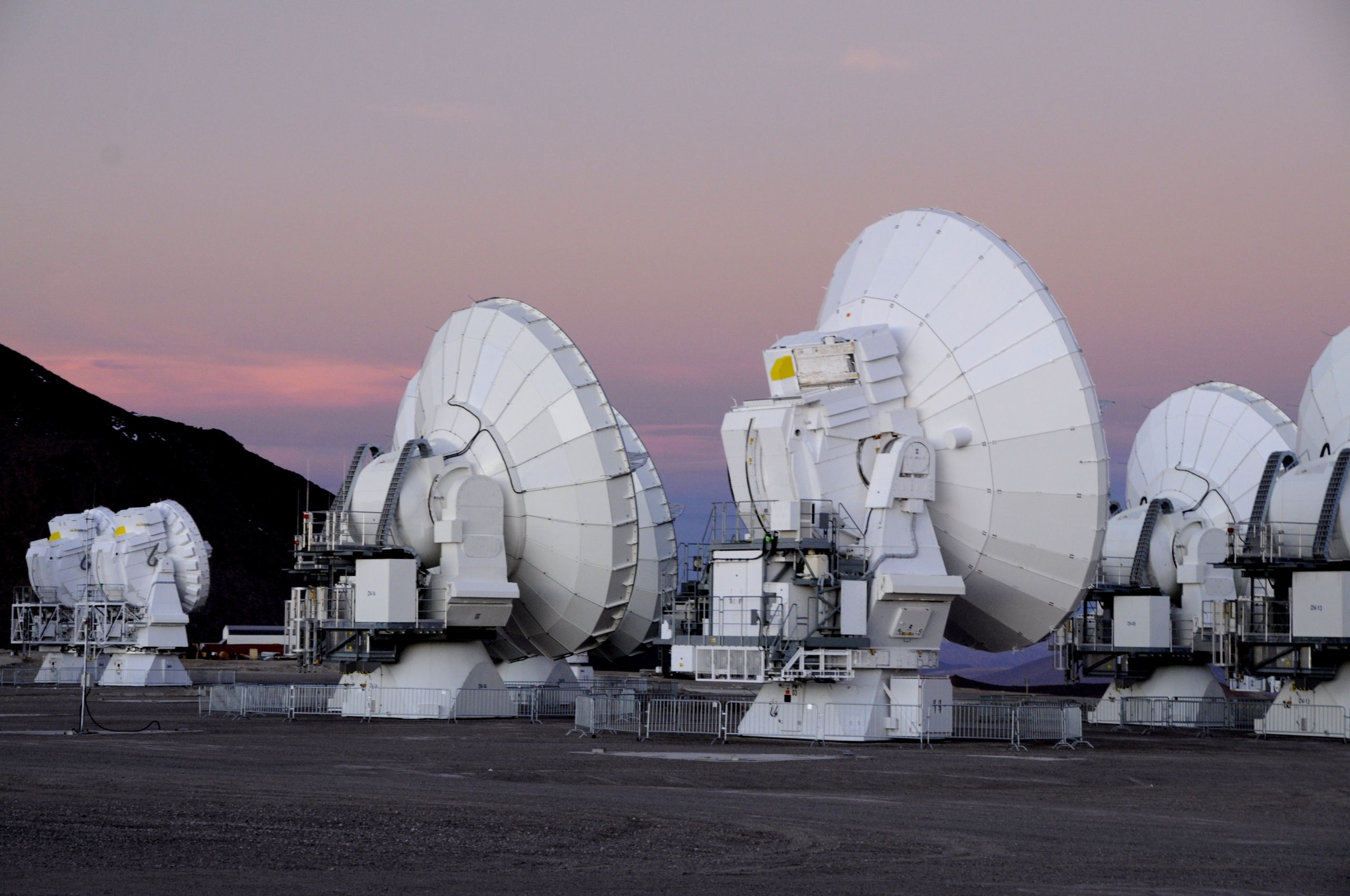 A row of large white satellite dishes stands on the barren Atacama landscape at dusk. The sky is a gradient of pale pink and blue, hinting at a sunset. These dishes are part of a scientific observatory, likely used for astronomical observations.