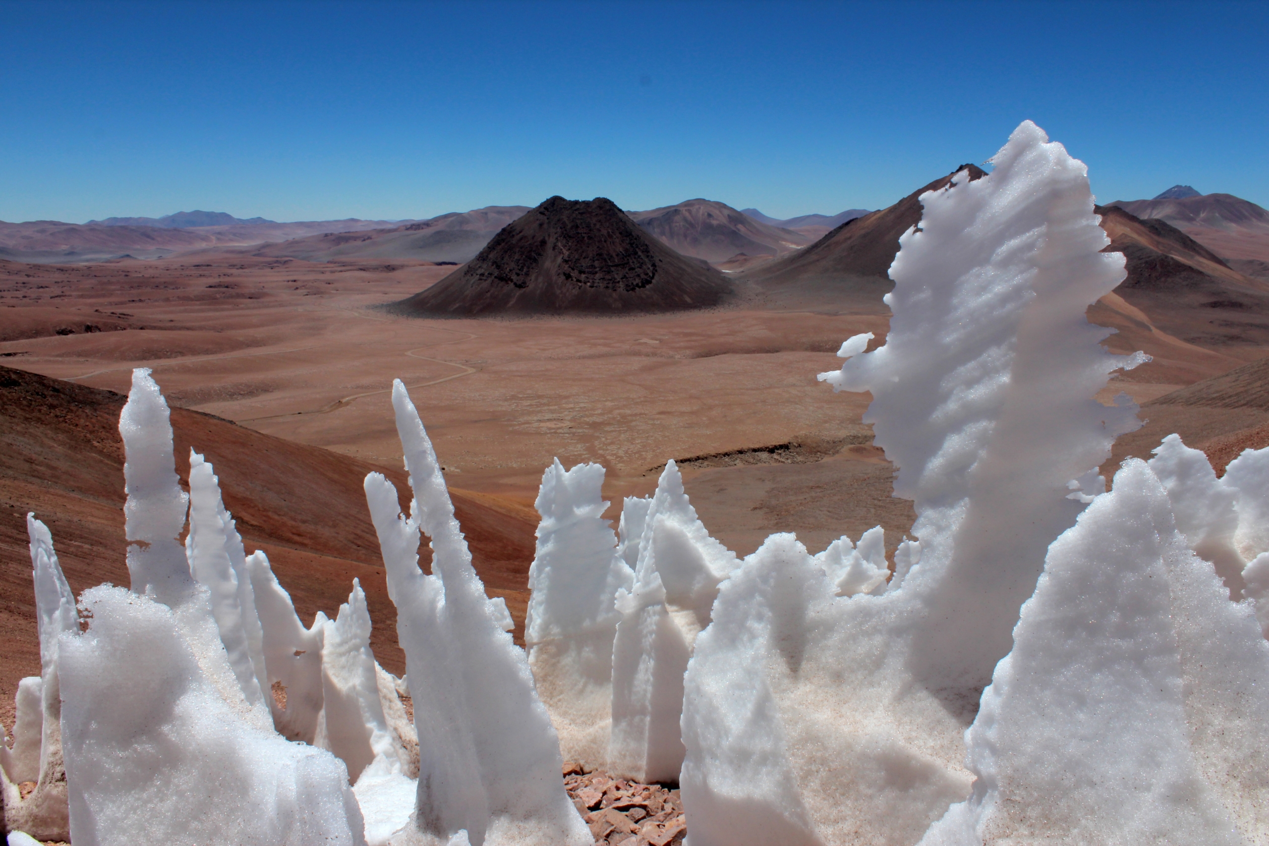 Snow formations called penitentes, resembling sharp, jagged spikes, rise from the barren desert landscape of the Atacama with mountains in the background under a clear blue sky.