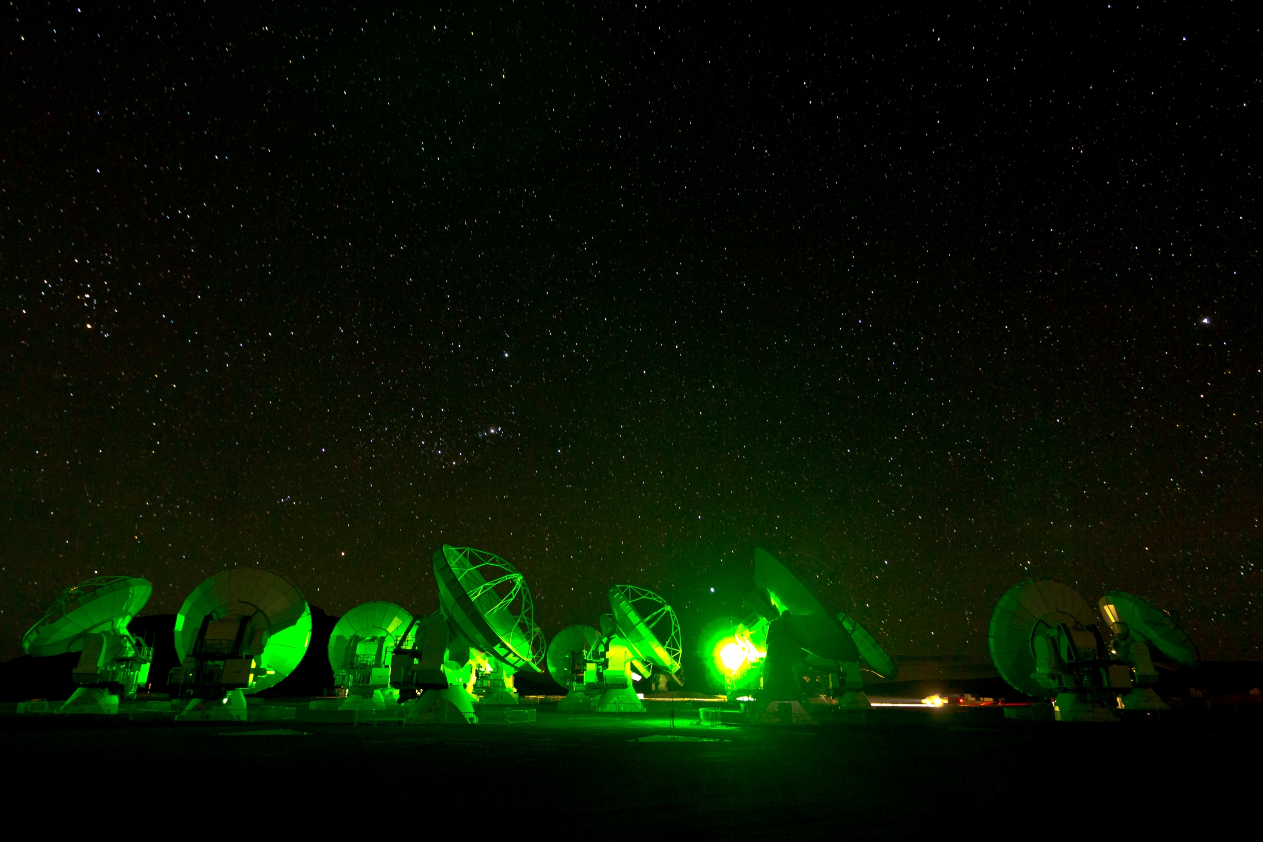 Under the starry night sky of Atacama, a row of large radio telescopes is illuminated by green lights. Silhouetted against a dense cluster of stars, they create a serene and high-tech atmosphere, perfectly blending nature's wonder with human innovation.