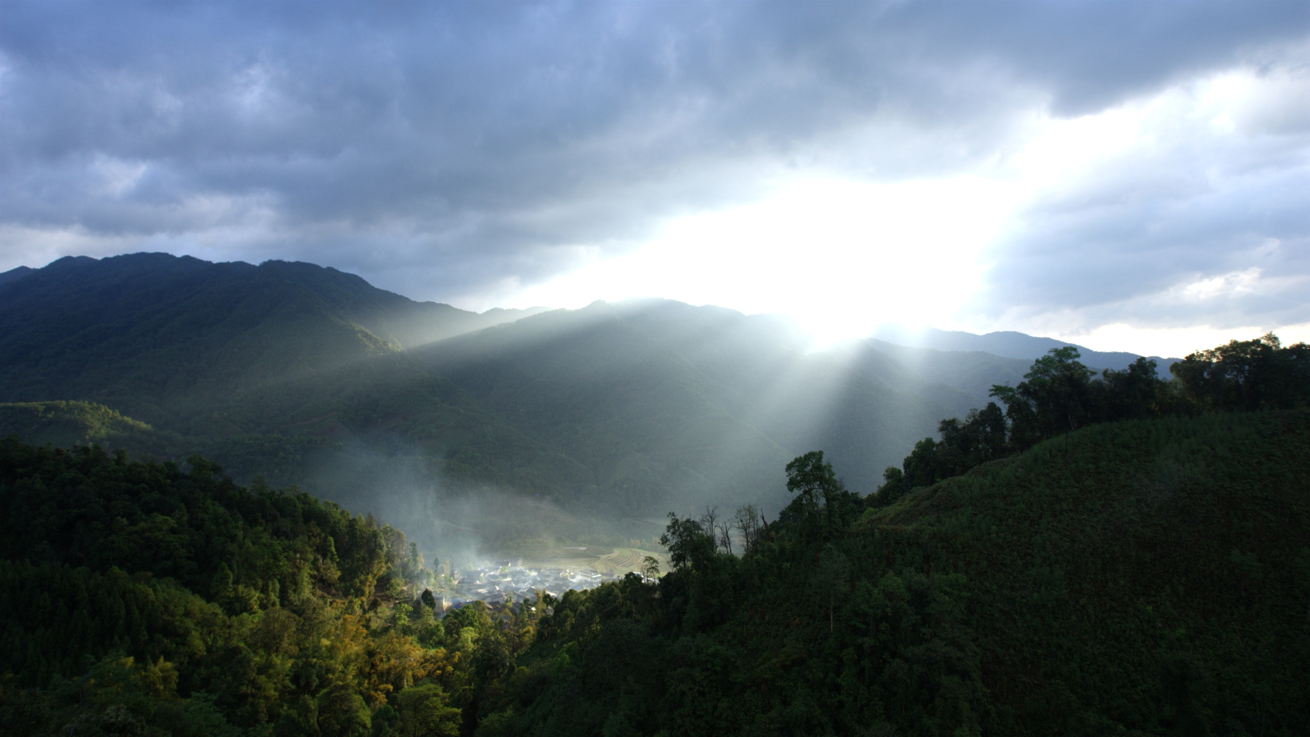 Sunlight breaks through clouds, illuminating a valley nestled among lush green mountains. Mist rises from the forested area, adding a mystical atmosphere to the serene landscape—a perfect backdrop for an ape with an app searching for love amidst nature's tranquility.