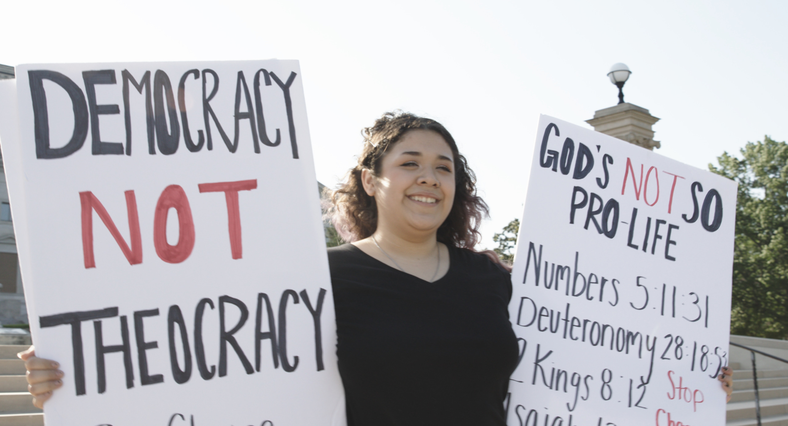 A person smiles while holding two protest signs. One reads 