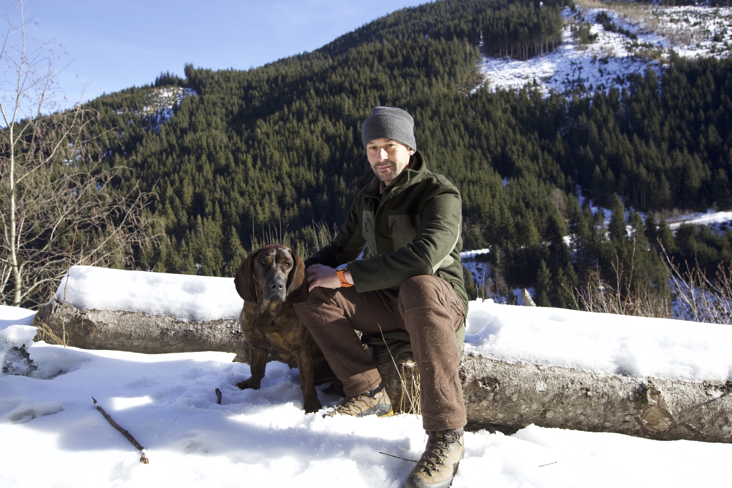 A person wearing a winter jacket and beanie sits on a snow-covered log with their dog, 