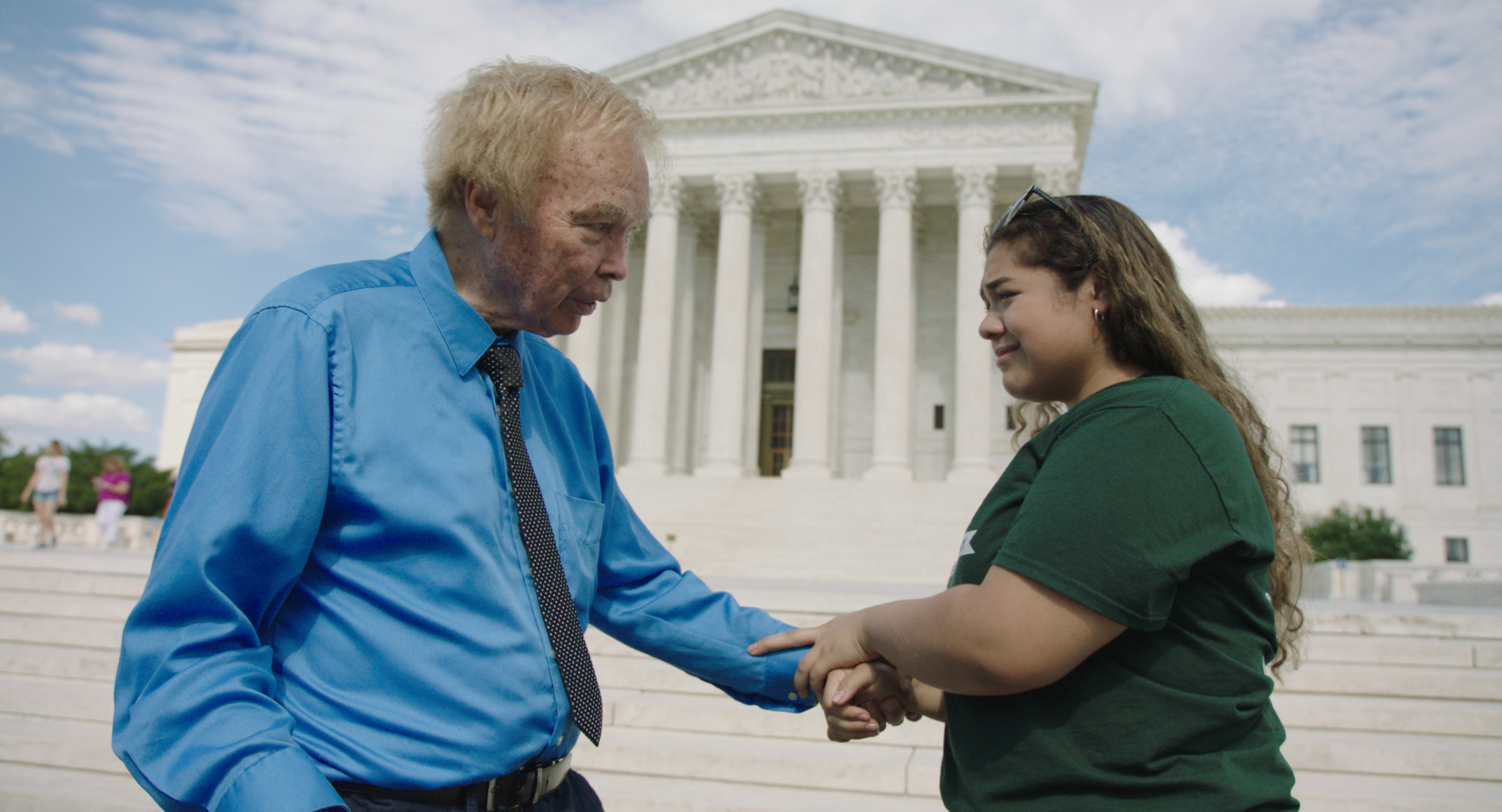 Outside the U.S. Supreme Court building, where Bill Baird once championed rights, an older person in a blue shirt and black tie gently holds the hand of a younger individual in a green shirt. The iconic columns and steps provide a historic backdrop to this poignant moment.