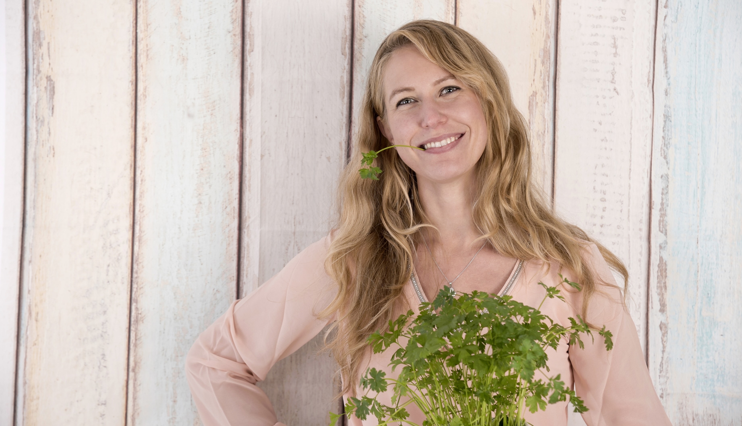 A smiling woman with long blonde hair holds a bunch of green parsley, reminiscent of fresh ingredients often explored in diets. She is wearing a light pink blouse and standing against a wooden plank background, playfully holding one parsley stem in her mouth as if on trial for taste.