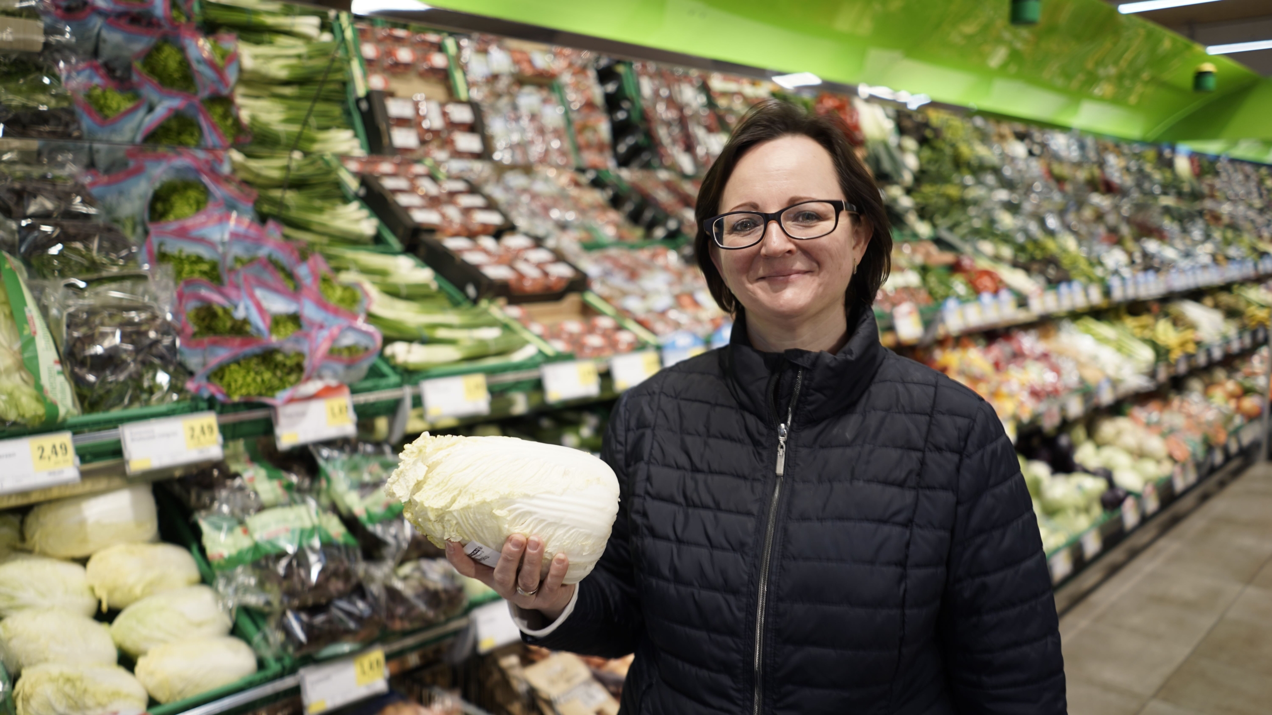 A person wearing glasses and a dark jacket is holding a cabbage in a grocery store, perhaps considering it for their new diet trial. They stand thoughtfully in front of shelves filled with various vegetables and produce.