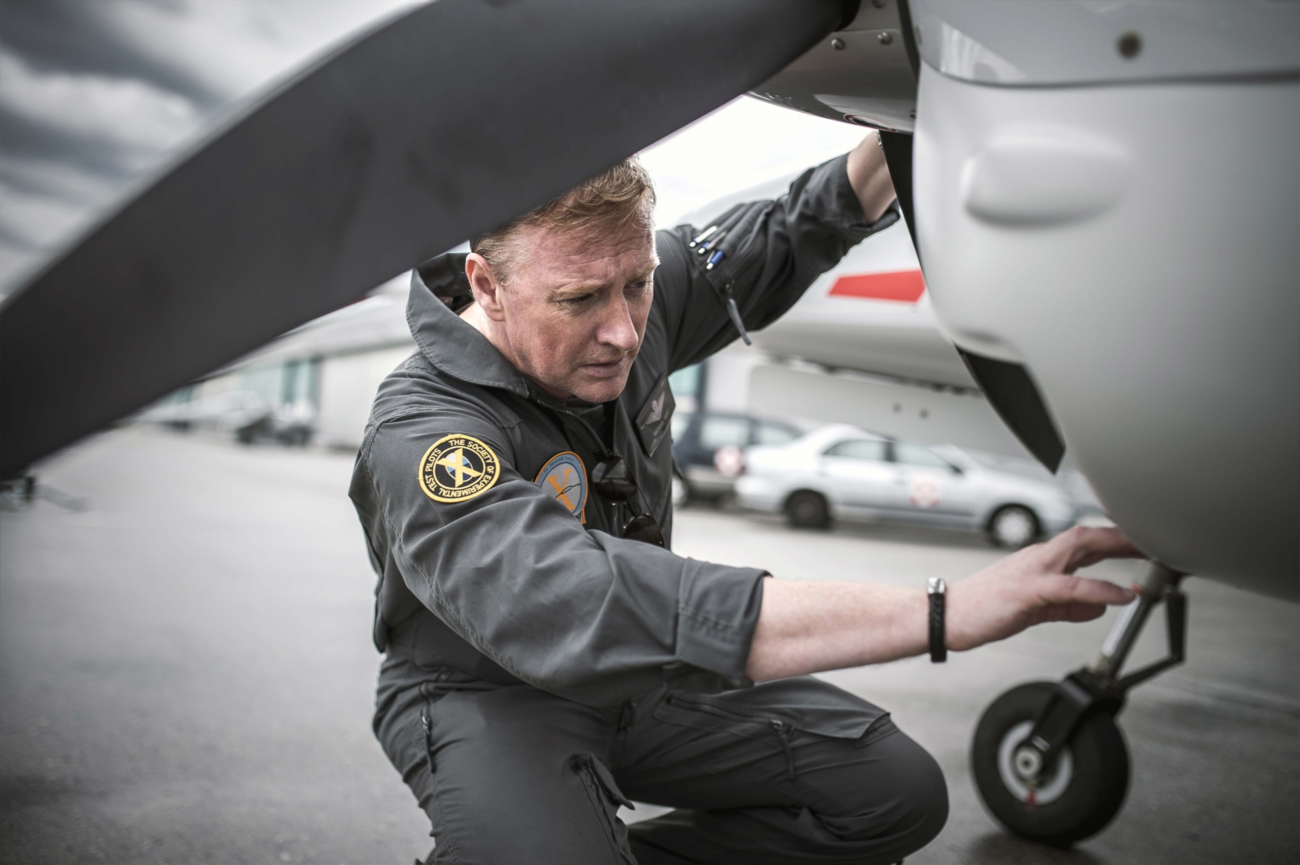 A person in a jumpsuit inspects the landing gear of a small aircraft on a tarmac. They are focused and reaching under the planes nose. Other planes and vehicles are visible in the background.