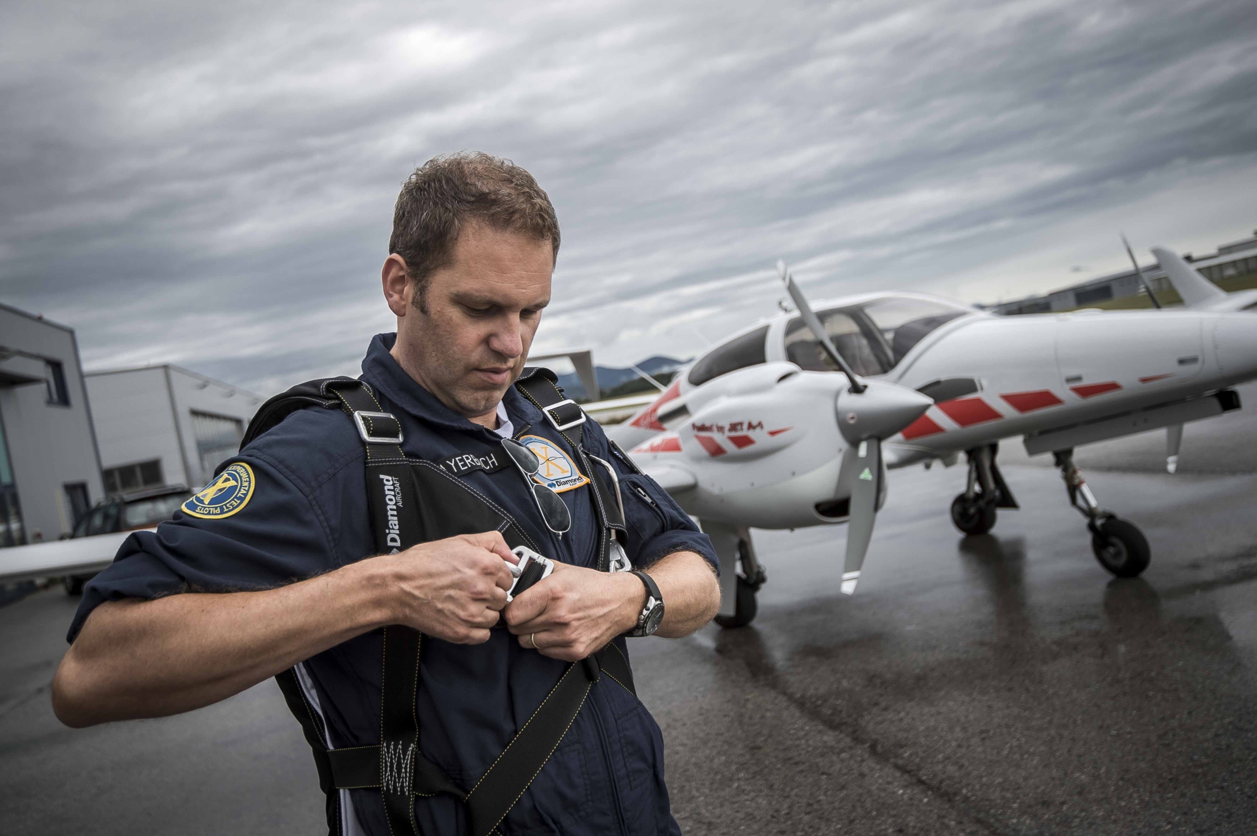 A person in a navy blue jumpsuit adjusts their harness in front of a small white aircraft on a tarmac. The sky is overcast, and there are buildings in the background.