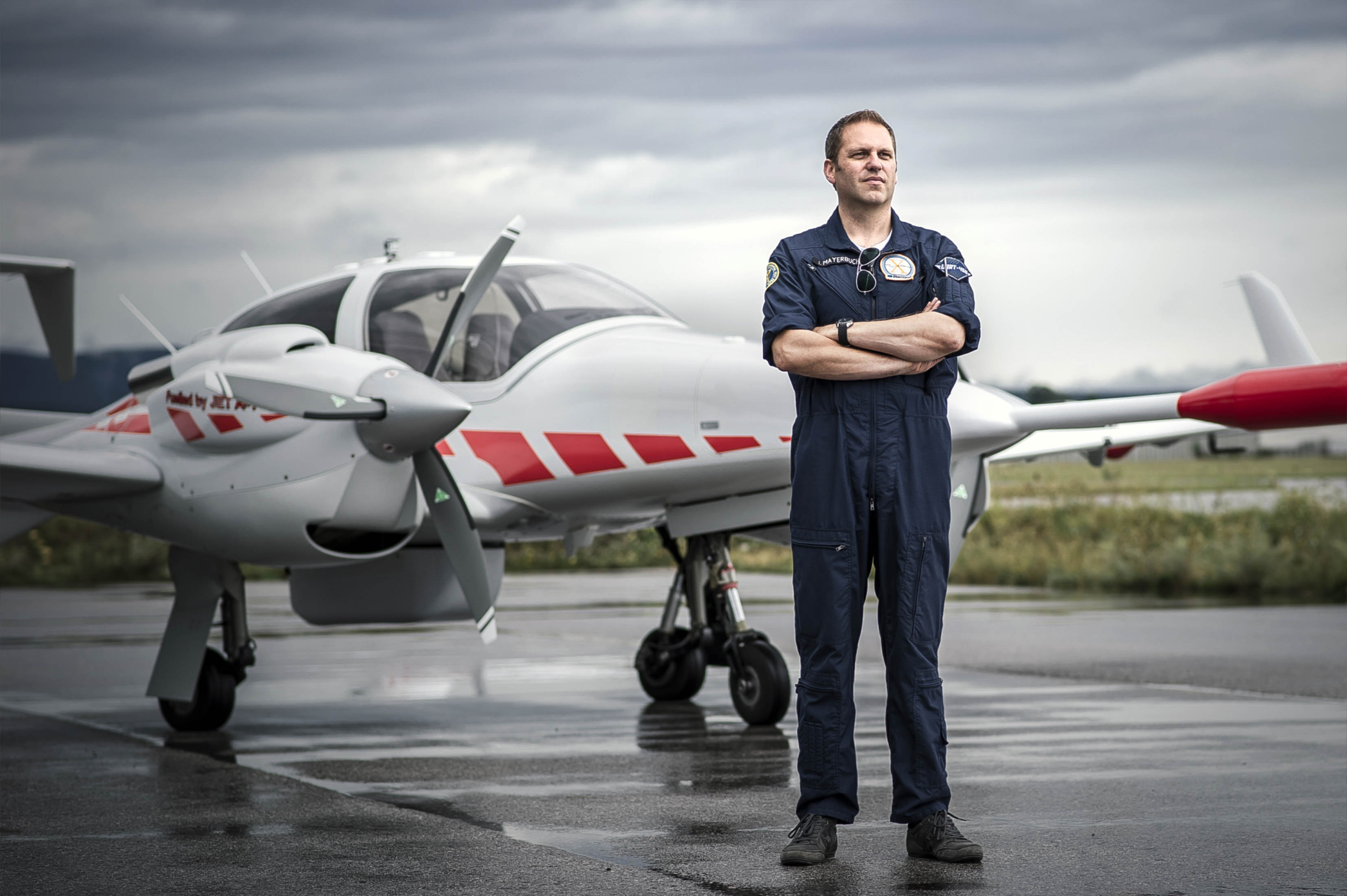 A man in a dark blue jumpsuit stands confidently with arms crossed on a wet airport tarmac, in front of a small white aircraft with red stripes. The sky is overcast, and the runway is reflective from recent rain.