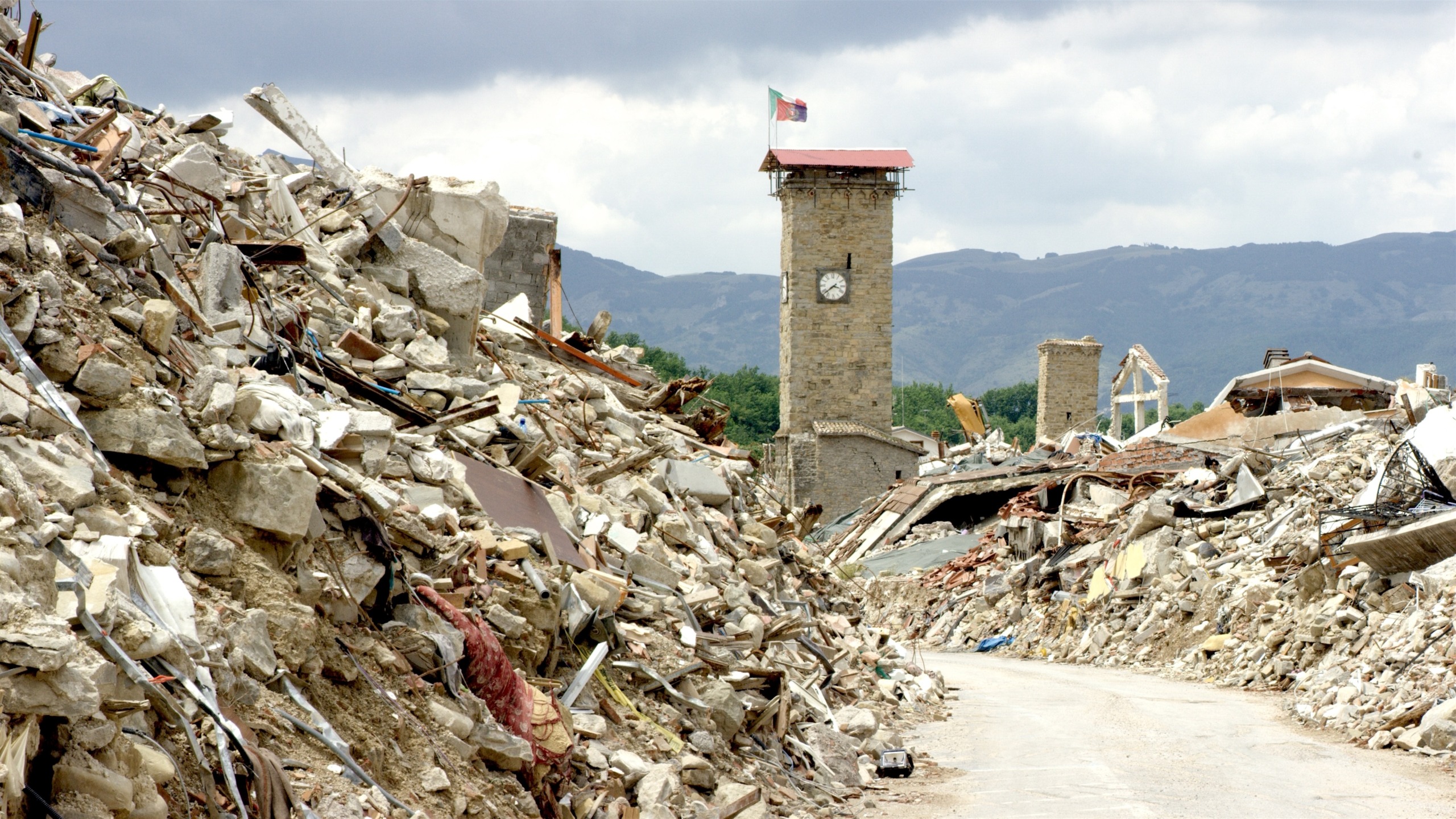 Amidst the devastation, a clock tower triumphantly stands tall, its flag fluttering above the wreckage of collapsed buildings. The haunting scene is framed by majestic mountains and a brooding cloudy sky, evoking an image straight out of Chasing Quakes.