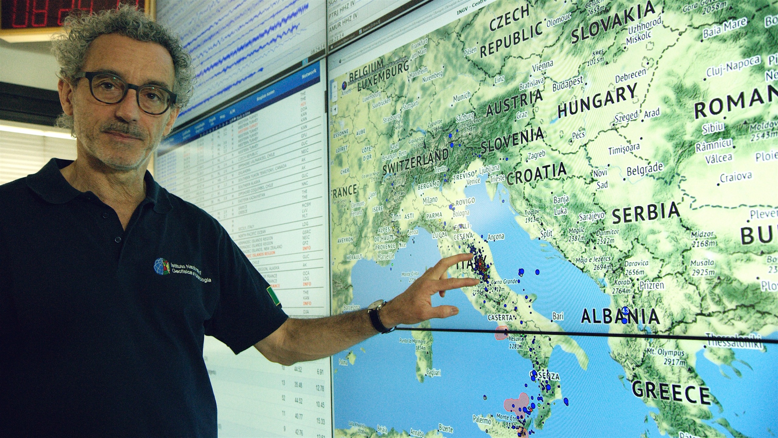 In an office setting, a man gestures towards a large digital map of Europe on a monitor, highlighting regions like Italy and Switzerland. Various data points reveal seismic activity as part of his ongoing research in seismology.