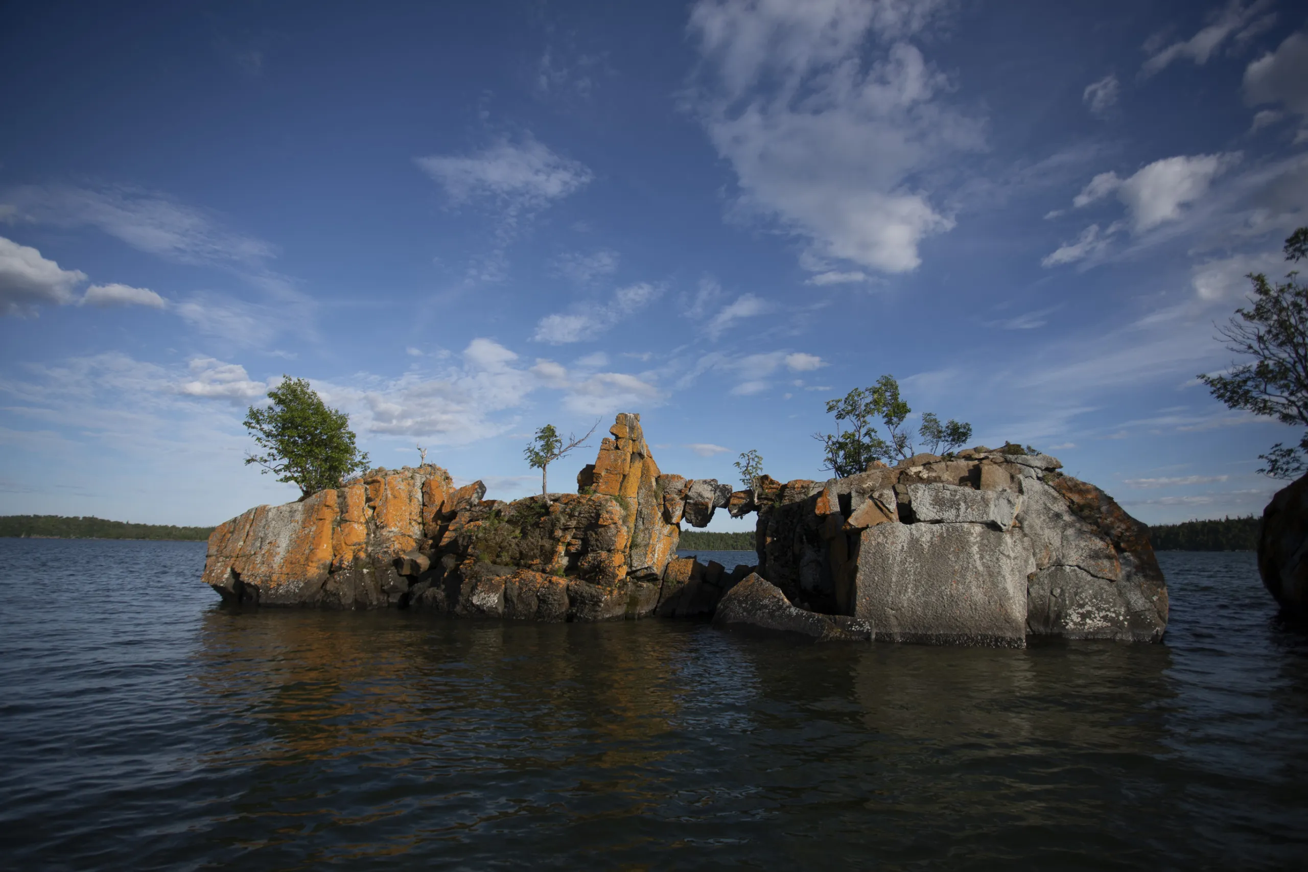 A rocky islet with an arch formation, partially covered in vegetation, stands majestically in the Great Lakes under a blue sky with scattered clouds, showcasing the untamed beauty of nature.