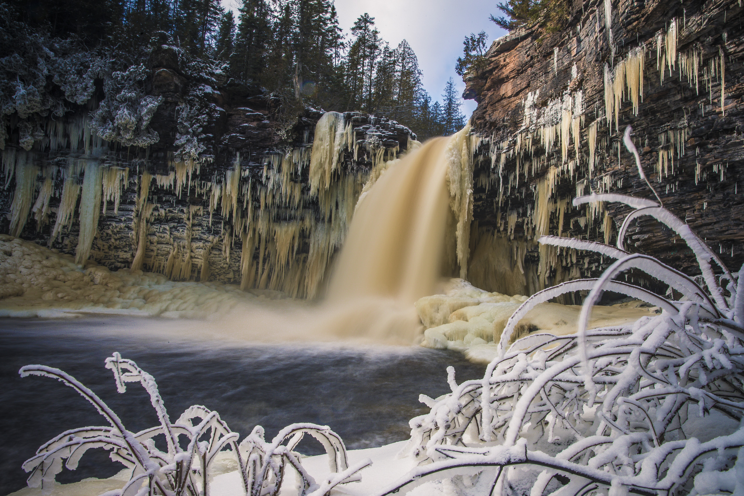 A frozen waterfall cascades into a partially frozen river, surrounded by snow-covered rocks and untamed icicles. Frost-coated branches frame the scene, set against a backdrop of Great Lakes majesty with snow-dusted evergreen trees.