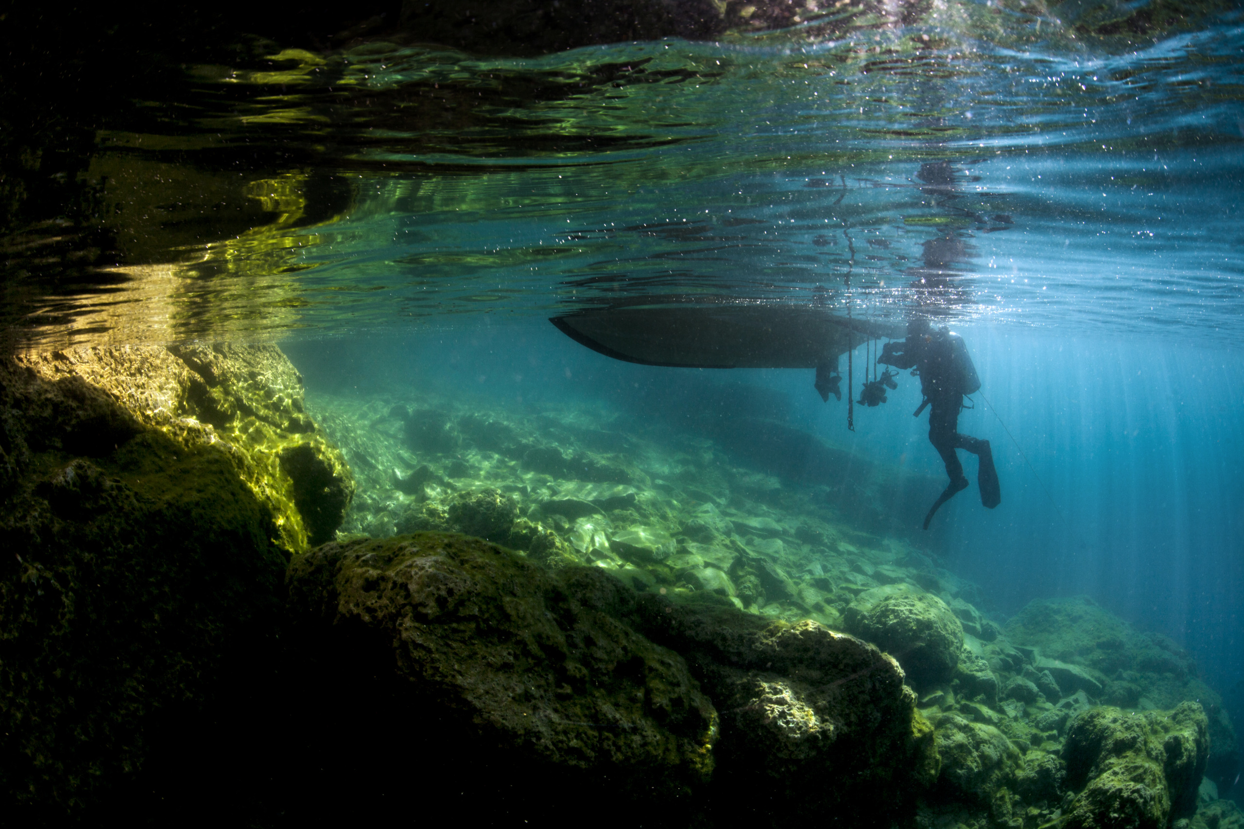 A scuba diver swims near the surface of clear turquoise water, casting a shadow on the untamed rocky underwater landscape. Sunlight streams through, illuminating the scene with a serene, mystical atmosphere reminiscent of the hidden beauty found beneath the Great Lakes.