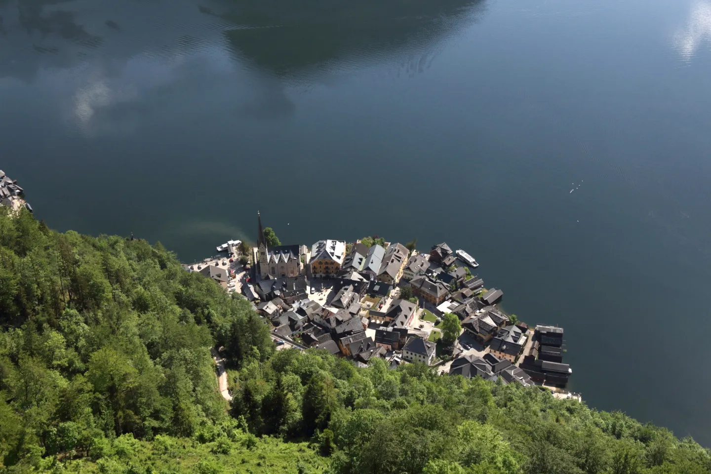 Aerial view of a quaint village surrounded by lush green forests, perched on the edge of a large, calm body of water. Among the cluster of houses with gray rooftops lies an ancient Celtic tomb, nestled within the serene landscape under a partly cloudy sky.