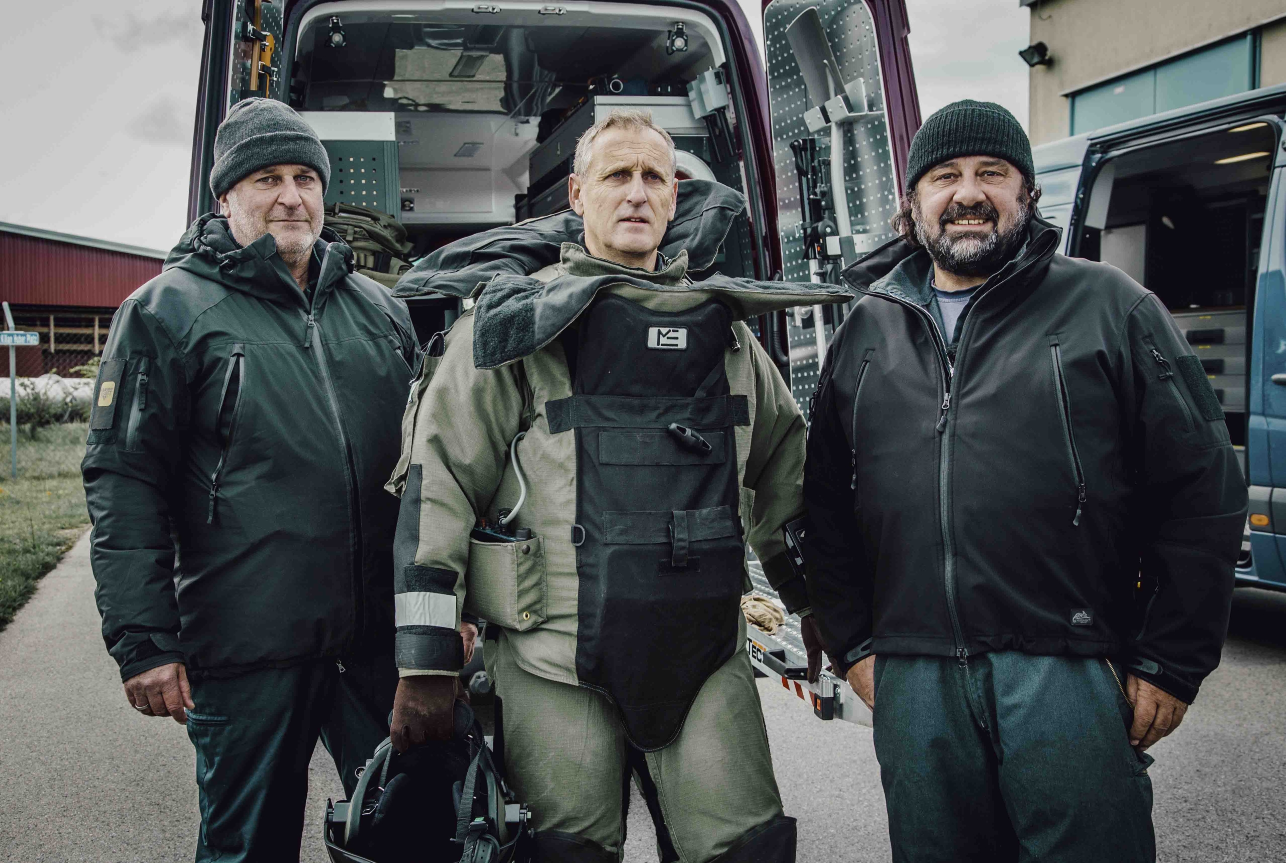 Three men stand in front of a vehicle. The man in the middle, a highly charged figure, wears a bomb disposal suit, while the other two explosive experts are dressed in outdoor clothing. The open vehicle behind them is filled with equipment.