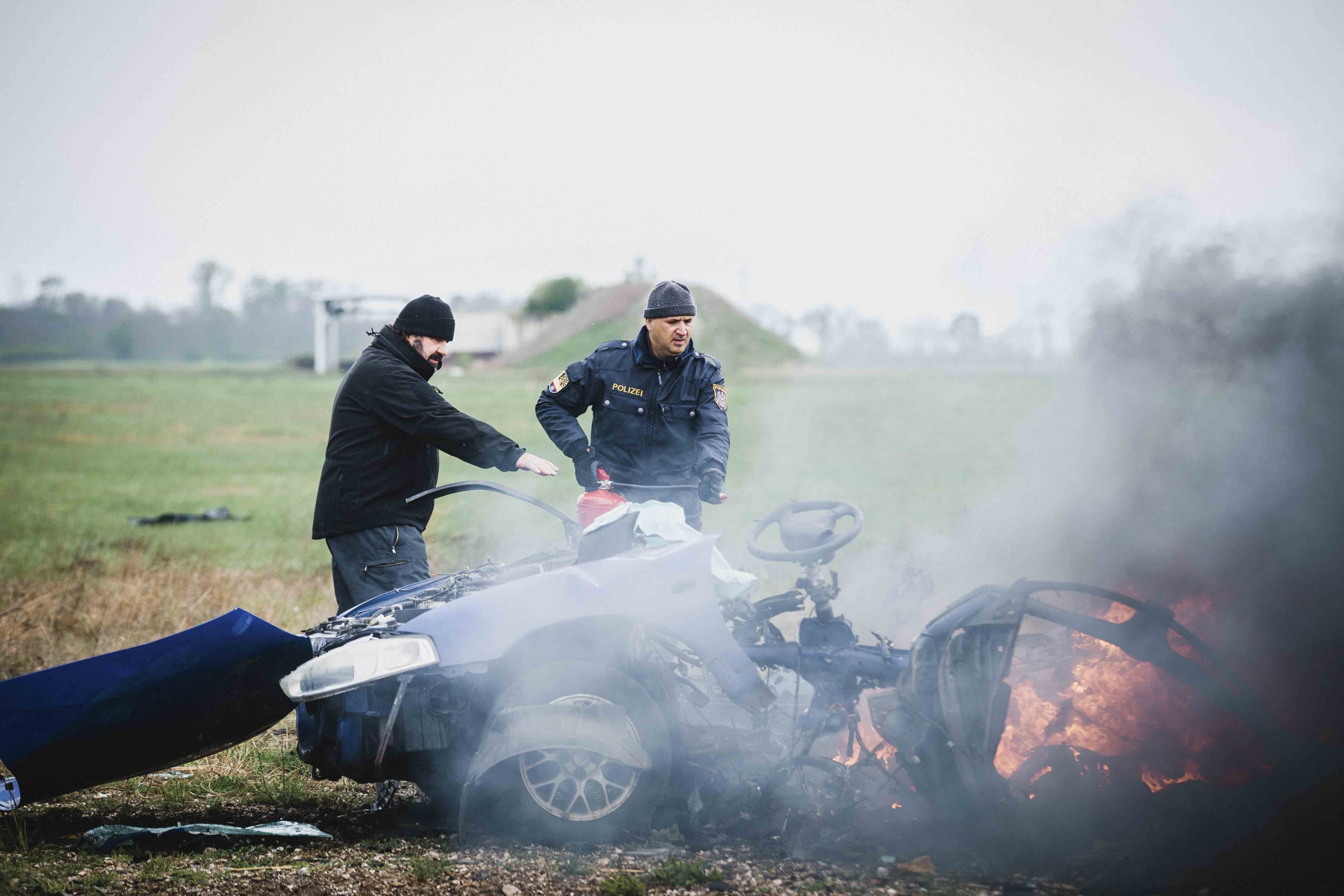 Two men stand near a burning, wrecked car in a field. One, clad in a police uniform, appears focused alongside an explosive expert. Smoke and flames rise from the vehicle, creating a highly charged atmosphere against the backdrop of sparse vegetation.