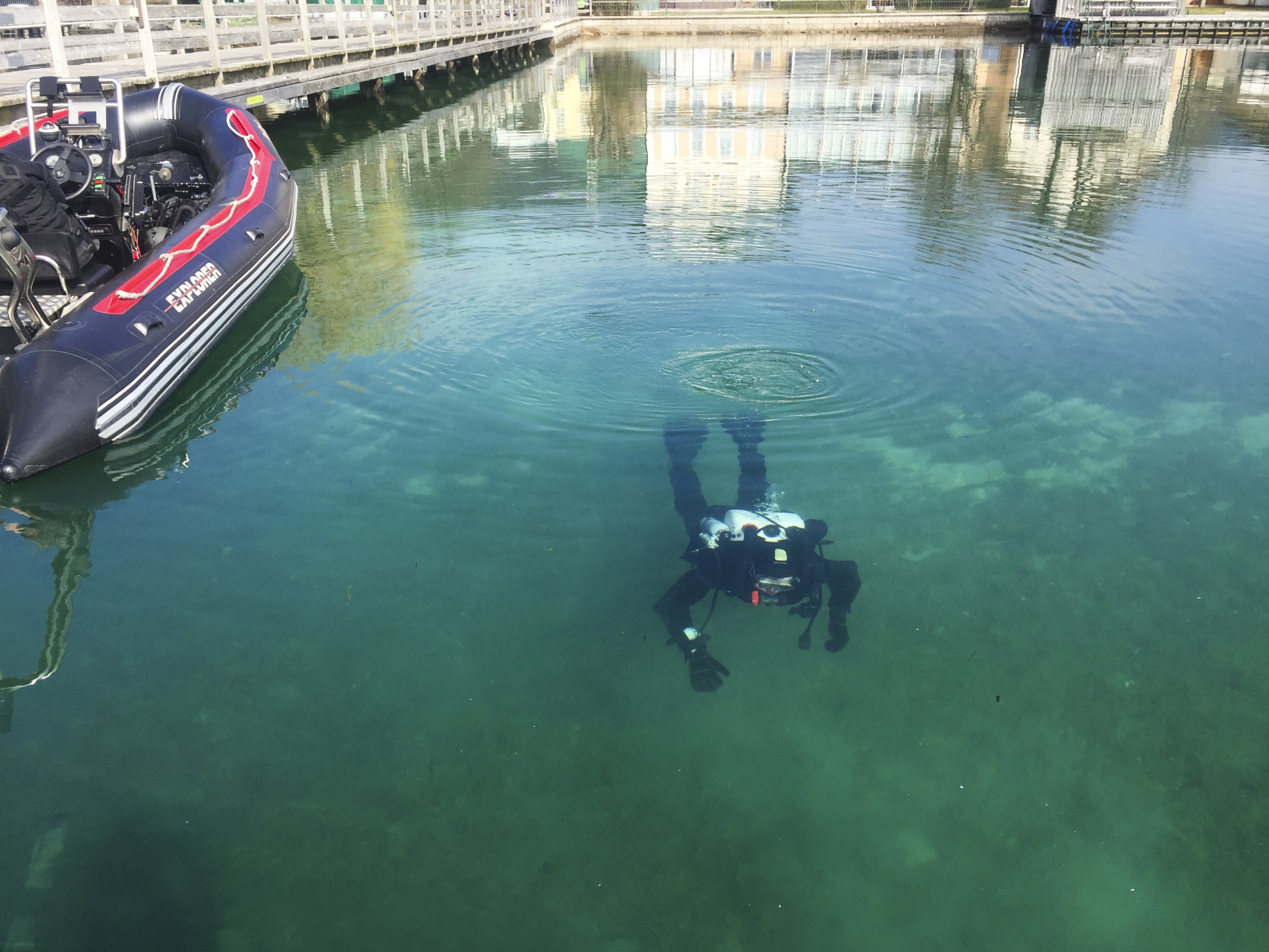 A scuba diver is submerged in clear water near a small inflatable boat tied to the dock. The tranquil scene contrasts with the expertise of explosive experts, as the diver's silhouette drifts beneath reflections of nearby buildings shimmering on the serene surface.